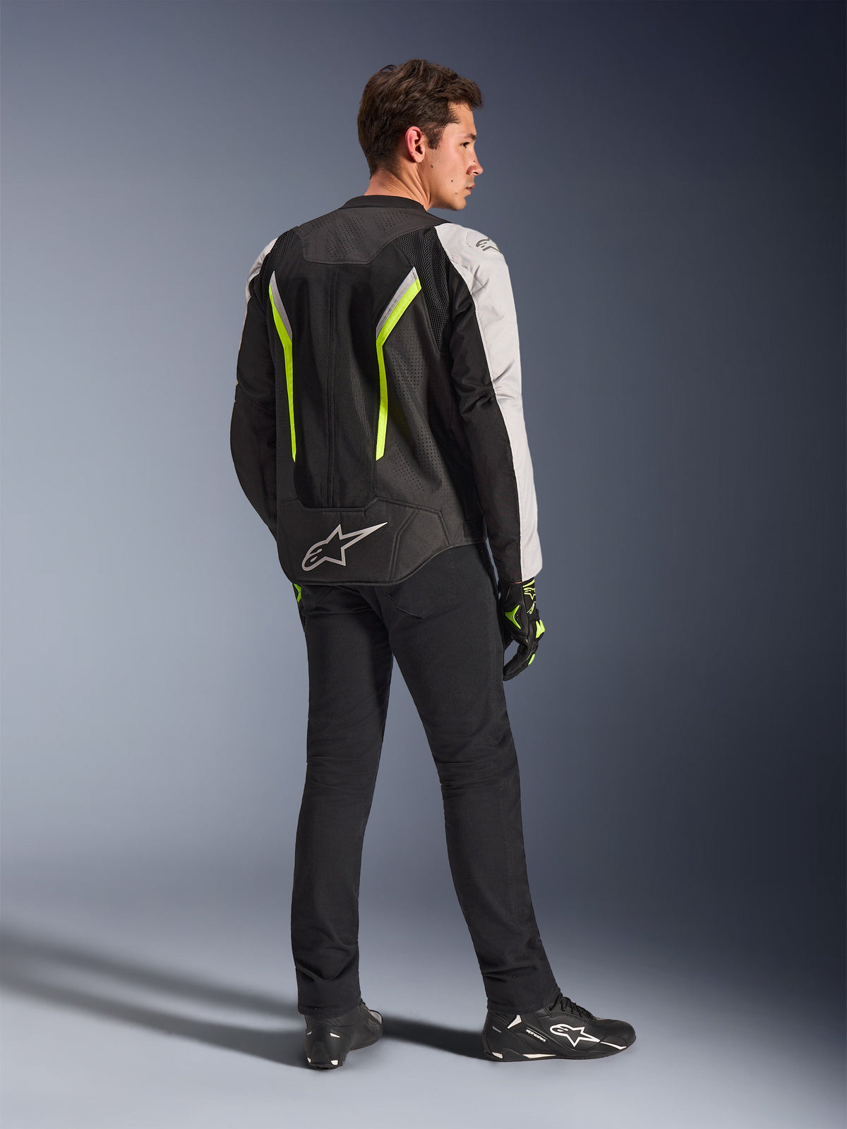 Alpinestars AST V3 Air Jacket in Black Silver Yellow Fluo, textiele motorjas gedragen door een mannelijke rijder vanaf de achterkant gezien, met uitgebreide mesh-panelen en fluorescerende gele accenten, gecombineerd met een zwarte motorbroek, zwarte handschoenen en zwarte Alpinestars schoenen, toont technische uitrusting voor sportief rijden.
