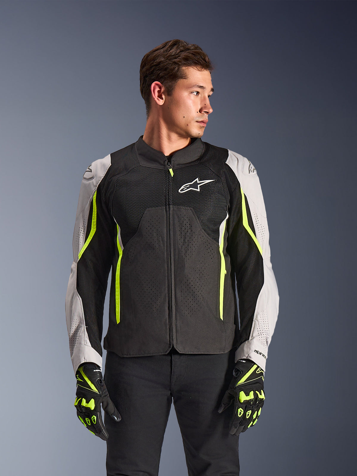 Alpinestars AST V3 Air Jacket, textiel motorjas, zwart en zilver met geel fluo accenten, voorzien van grote mesh ventilatiepanelen en laserperforaties, gedragen door een model met bijpassende handschoenen, sportieve motoruitrusting