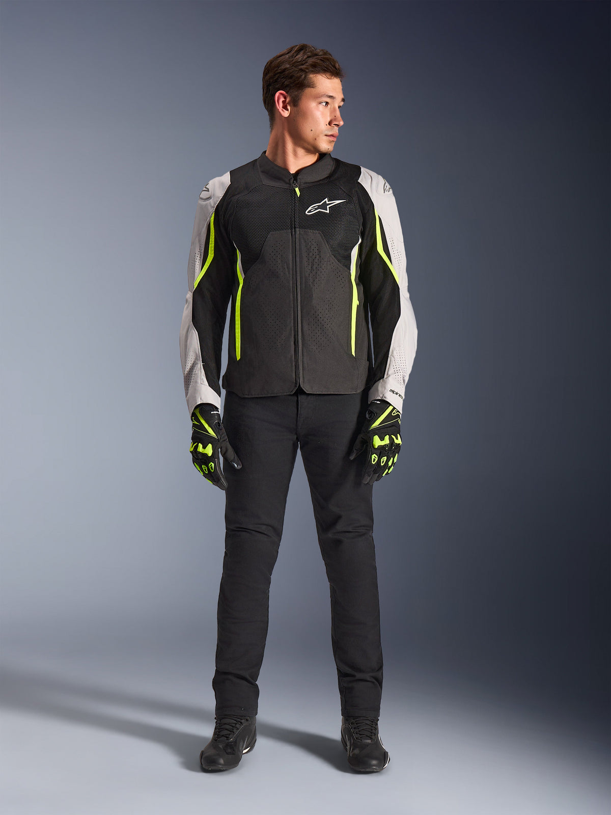 Mannelijk model in Alpinestars AST V3 Air Jacket in Black Silver Yellow Fluo, voorzien van mesh ventilatiepanelen en fluorescerende accenten, gecombineerd met een zwarte motorbroek en bijpassende motorhandschoenen, sportieve performance motoruitrusting.