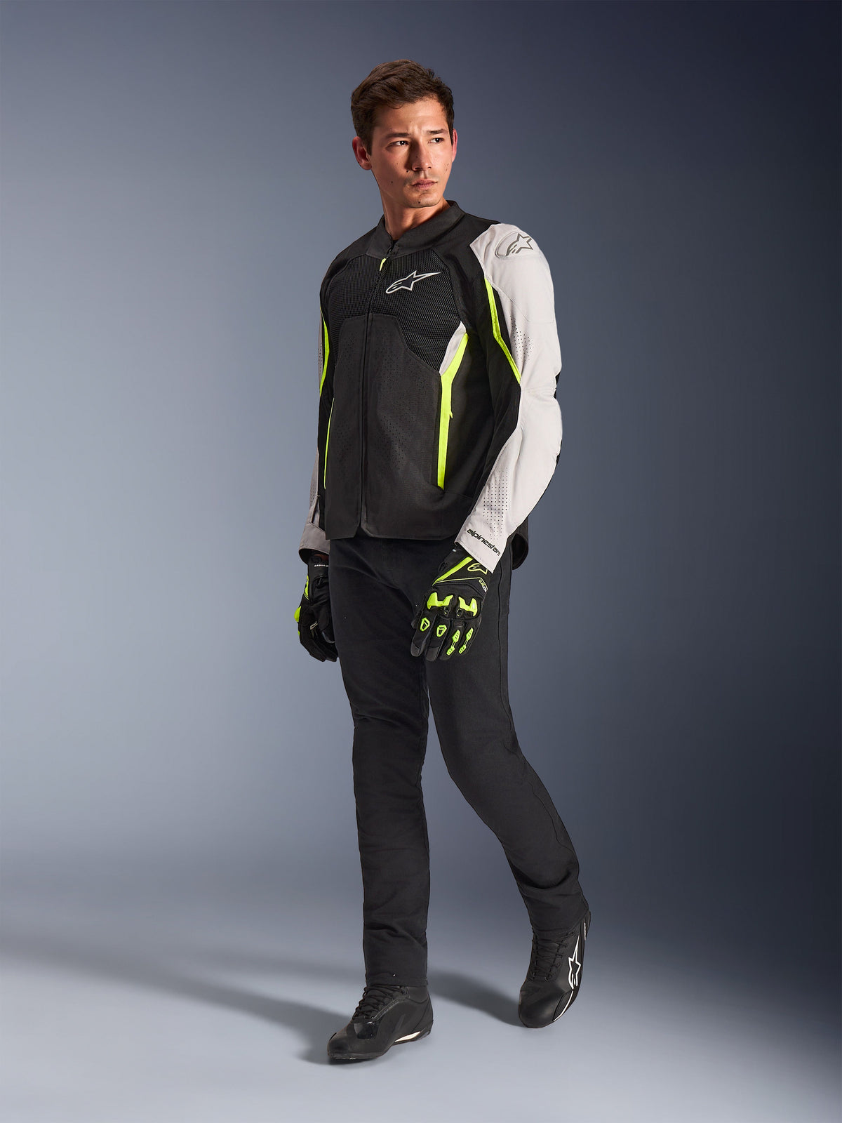 Alpinestars AST V3 Air Jacket, lichtgewicht textielen motorjas in zwart, zilver en Yellow Fluo, gedragen door een model met zwarte broek en bijpassende handschoenen, voorzien van mesh-panelen en laserperforaties voor sportief rijden bij warm weer.
