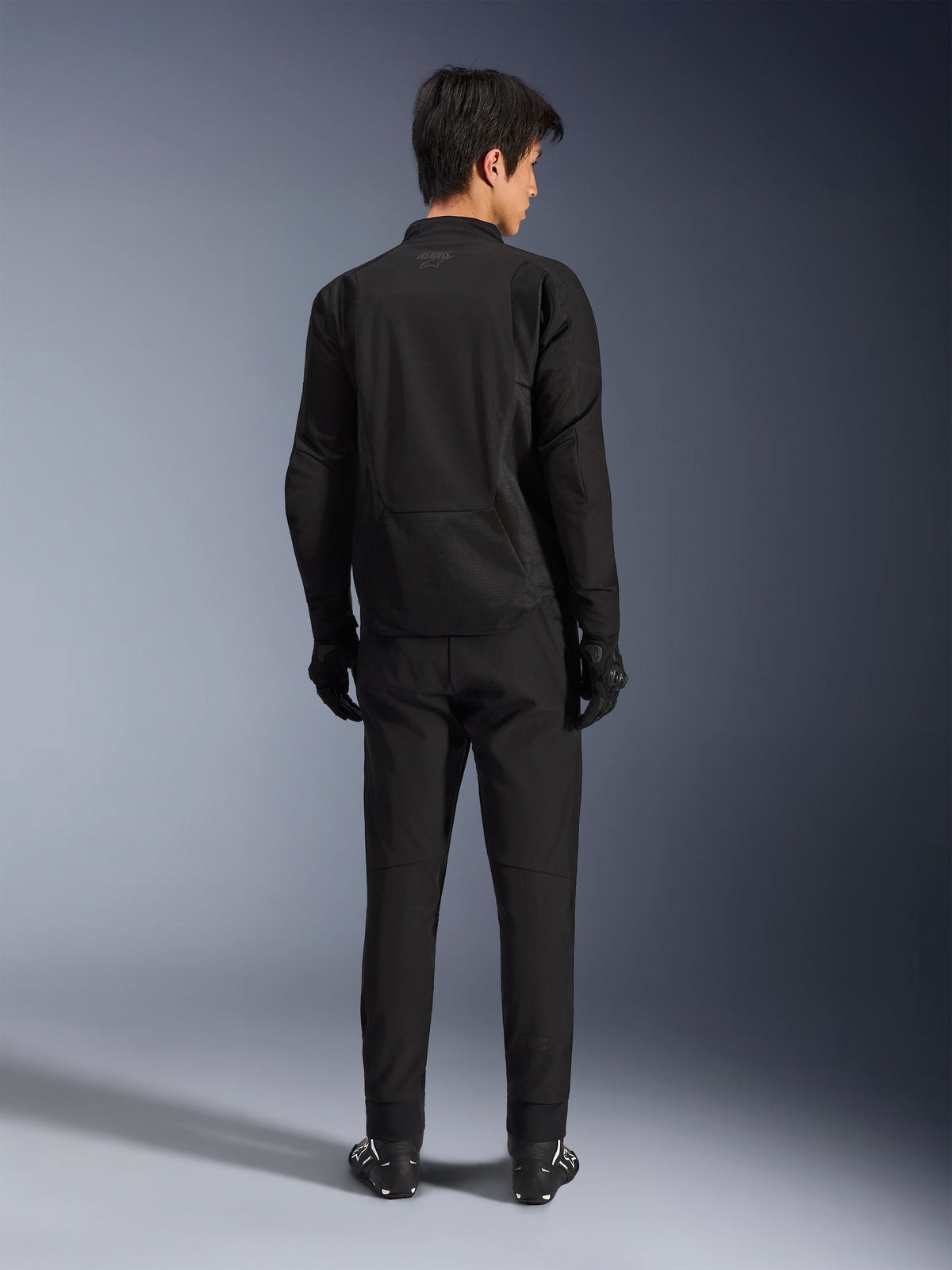 Alpinestars Aeroshell Airflow Jacket in Black Anthracite, rugaanzicht van een mannelijk model in volledige motoruitrusting inclusief zwarte broek en handschoenen, voorzien van een lichtgewicht softshell-constructie met een verlengde achterkant voor extra dekking.