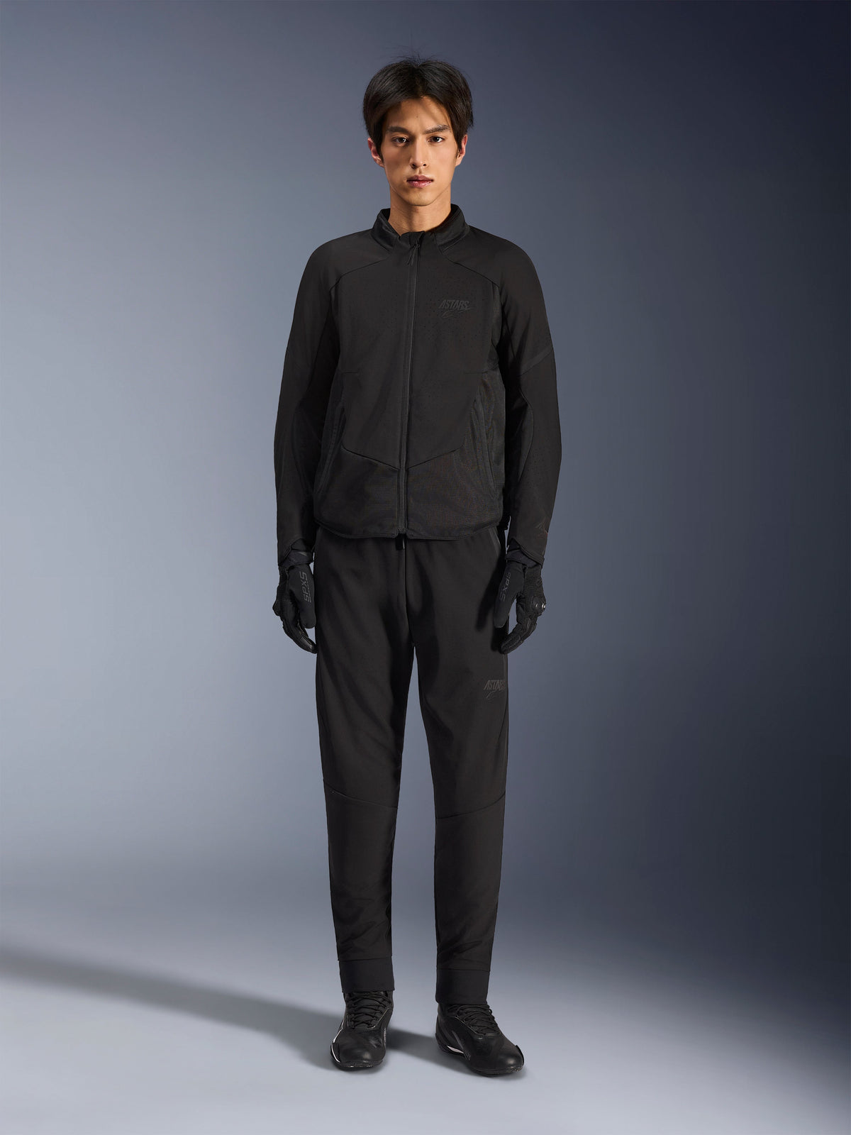 Alpinestars Aeroshell Airflow Jacket, zwarte motorjas, gedragen door model met bijpassende zwarte broek en handschoenen, voorzien van lasergeperforeerde borst- en zijpanelen van mesh, sportieve pasvorm voor prestaties bij warm weer