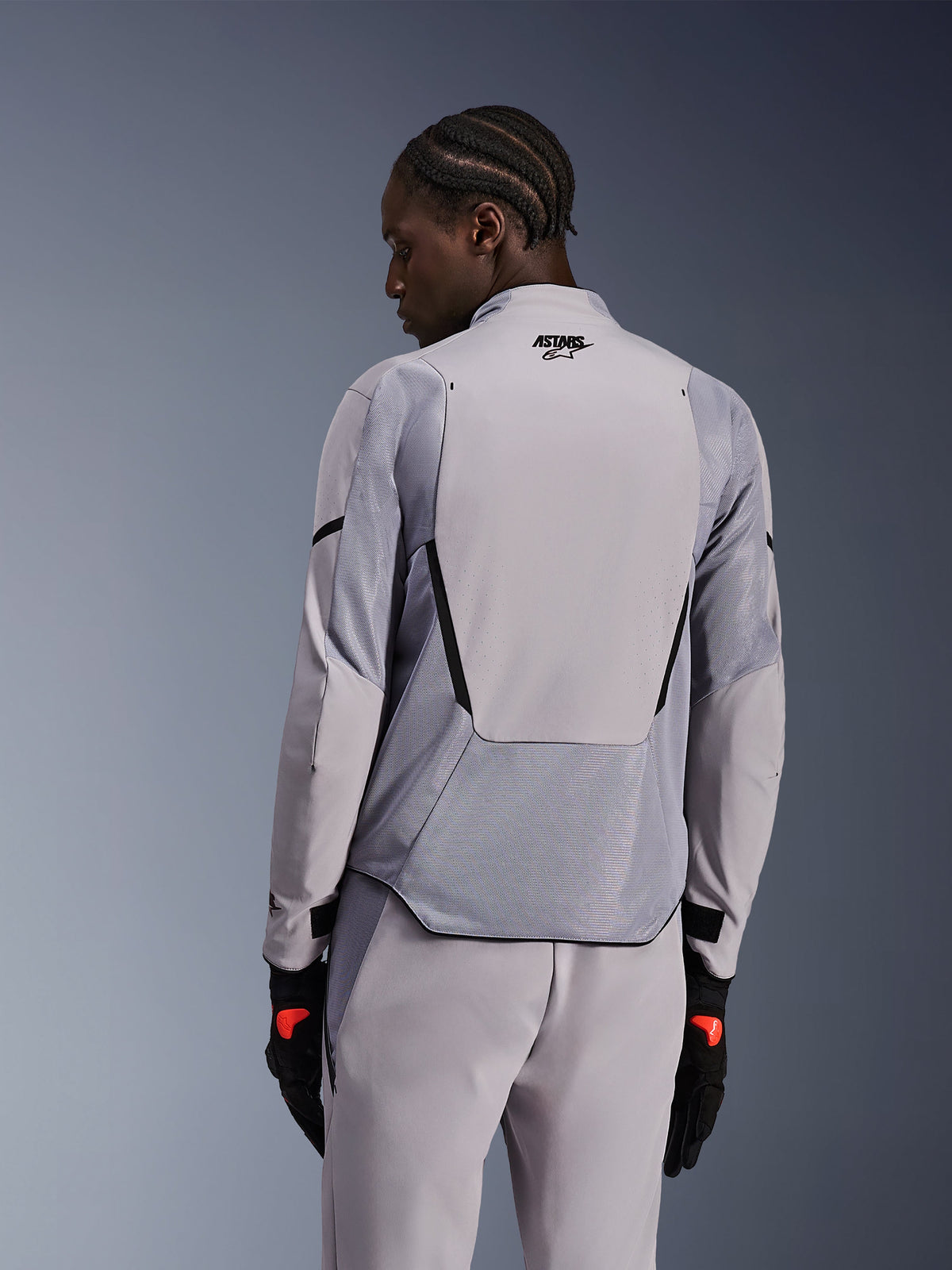 Alpinestars Aeroshell Airflow Jacket in Mid Gray Anthracite, achteraanzicht met ademende mesh-panelen en laserperforaties, technische stretch softshell-constructie met een verlengd rugpaneel, gedragen met bijpassende grijze broek en zwarte handschoenen voor een sportieve look