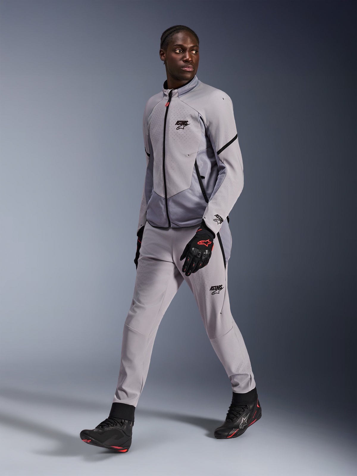 Alpinestars Aeroshell Airflow Jacket in Mid Gray Anthracite, gedragen door een mannelijk model met bijpassende grijze motorbroek en zwarte handschoenen, voorzien van laser-geperforeerde borstventilatie en een strak sportief ontwerp.