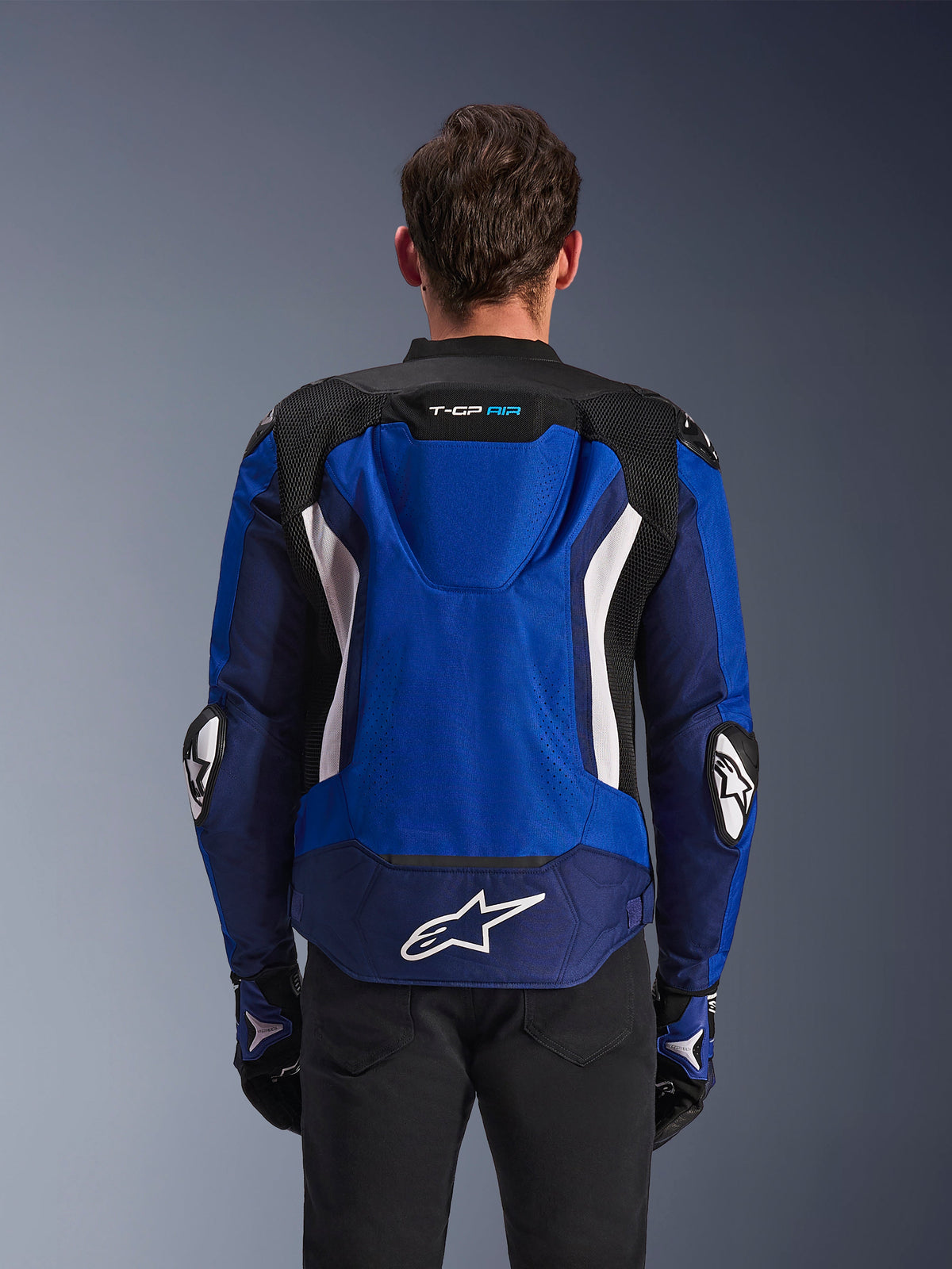 Alpinestars T-GP Air Jacket, sportieve textiele motorjas, helderblauw, navy en wit, achteraanzicht met de geïntegreerde aerodynamische bult en een groot wit logo op de onderrug, gedragen met bijpassende blauwe en zwarte motorhandschoenen