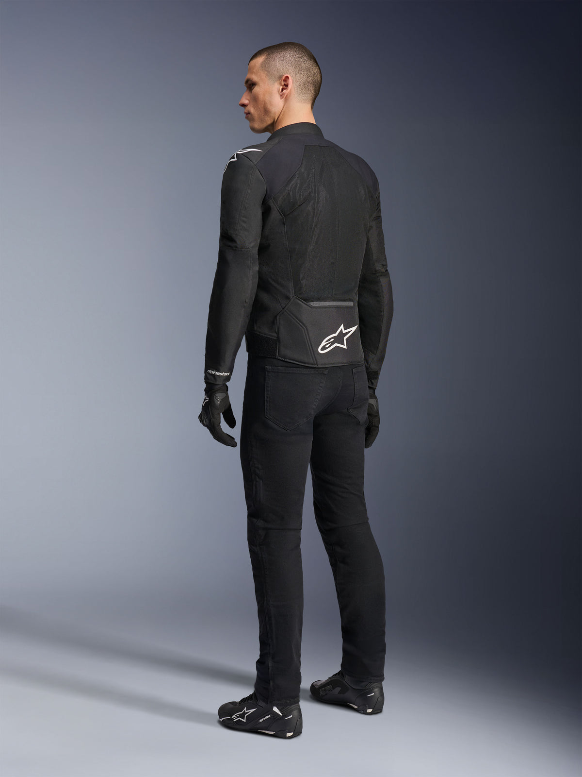 Alpinestars Viper Air V4 Jas, zwarte textiele motorjas, achteraanzicht op een mannelijk model, voorzien van grote tech mesh-panelen voor luchtstroom en een wit sterlogo op de onderrug, gedragen met bijpassende zwarte motorbroek en handschoenen