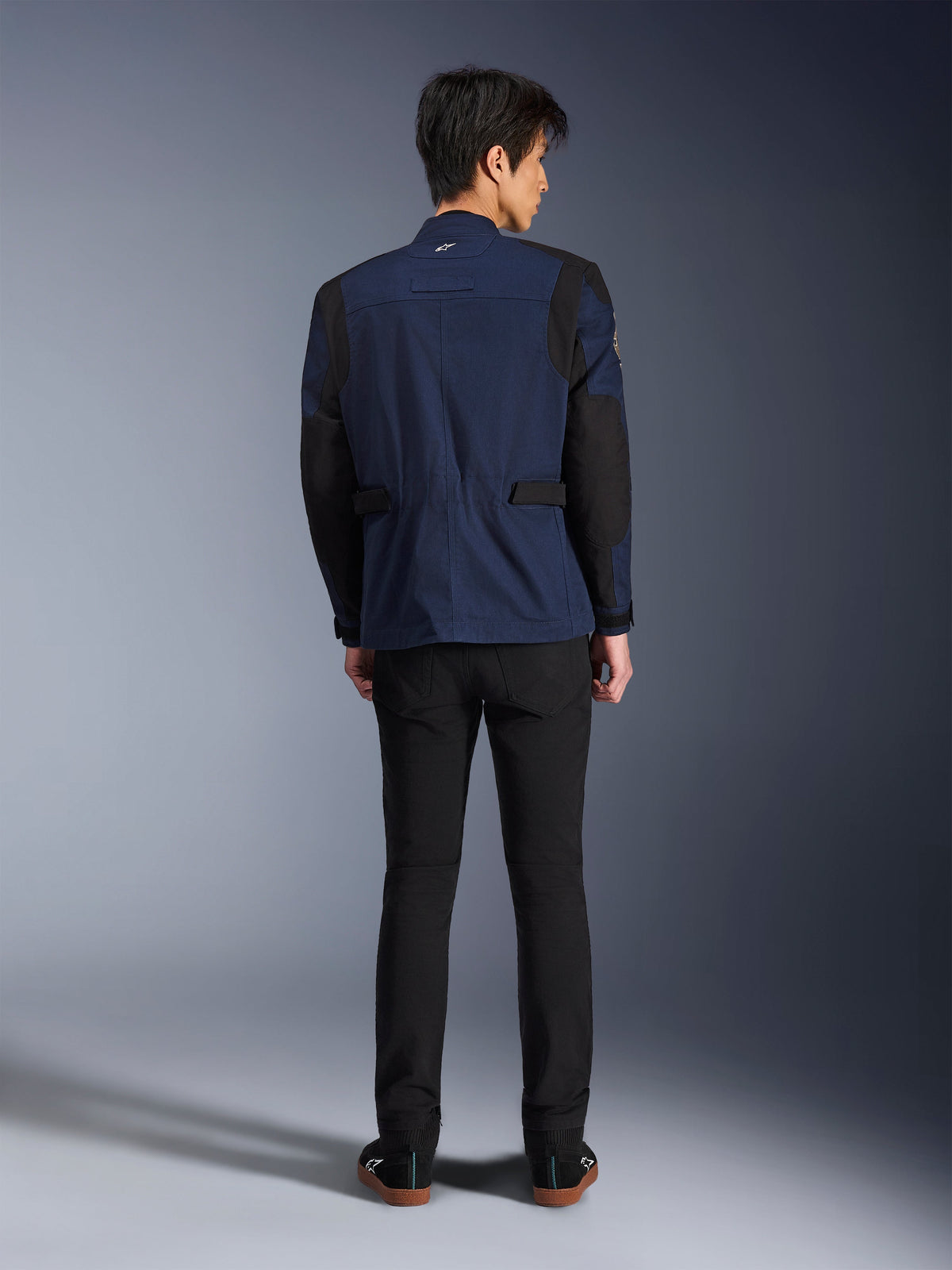 Model draagt de Alpinestars Dice Jacket in Dark Blue en Black, achteraanzicht, stedelijke motorveldjas met box-geplooide schouders en verstelbare tabs in de taille, gecombineerd met een zwarte broek en zwarte sneakers