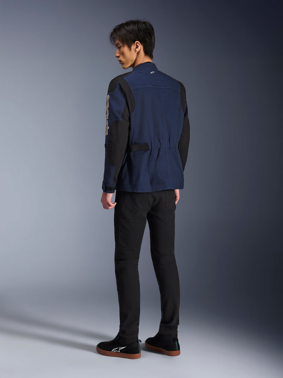 Mannelijk model van achteren gezien, draagt de Alpinestars Dice Jacket in donkerblauw en zwart, gecombineerd met een zwarte broek en zwarte sneakers, urban motorveldjas met geplooide schouders en verstelbare taille-tabs.