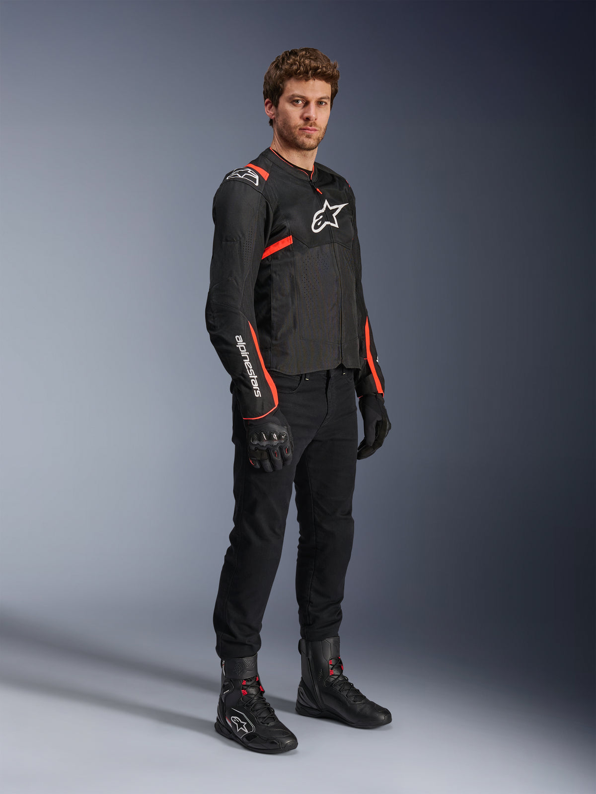 Mannelijk model in Alpinestars T-Sps Air v2 Jas, zwart met Red Fluo accenten, gecombineerd met een zwarte motorbroek, zwarte handschoenen en laarzen, sportief ontwerp met mesh-panelen en witte Alpinestars-logo's
