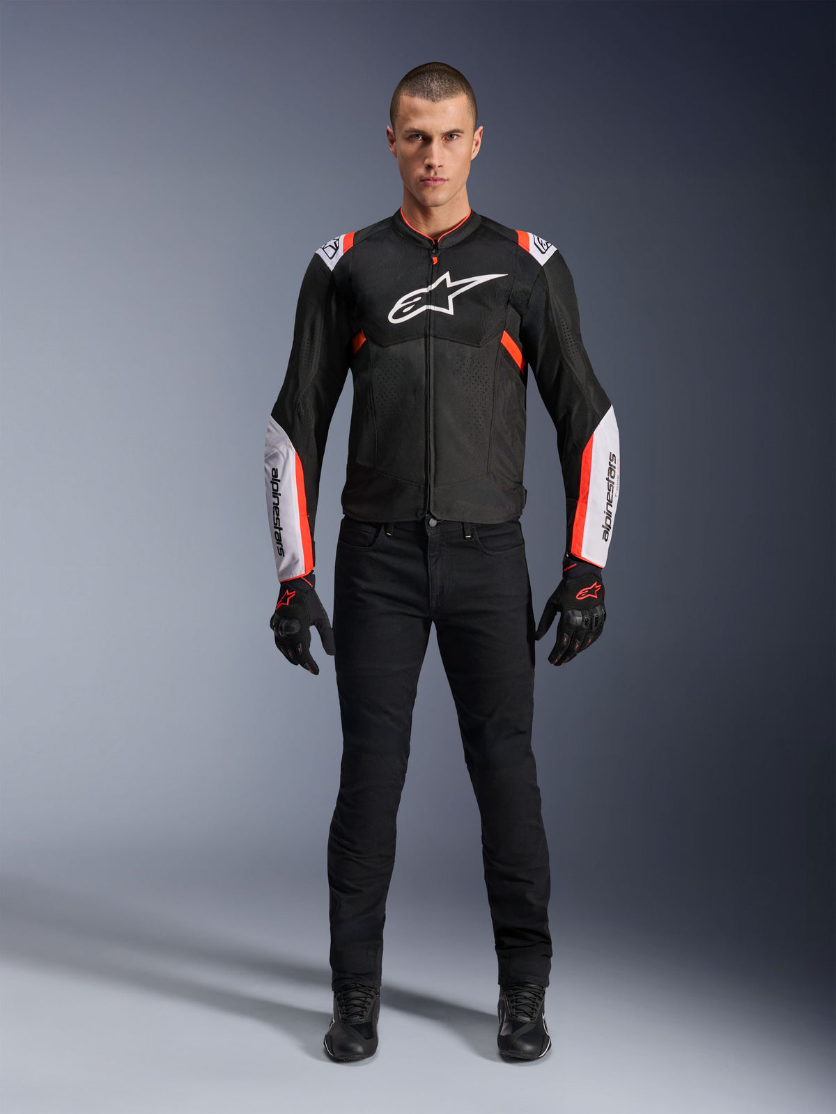 Mannelijk model draagt Alpinestars T-Sps Air V2 Jas in Black White Red Fluo, voorzien van grote tech mesh-panelen en opvallende witte logo's op de borst en mouwen, gecombineerd met een zwarte technische motorbroek en handschoenen, urban sport motorkleding