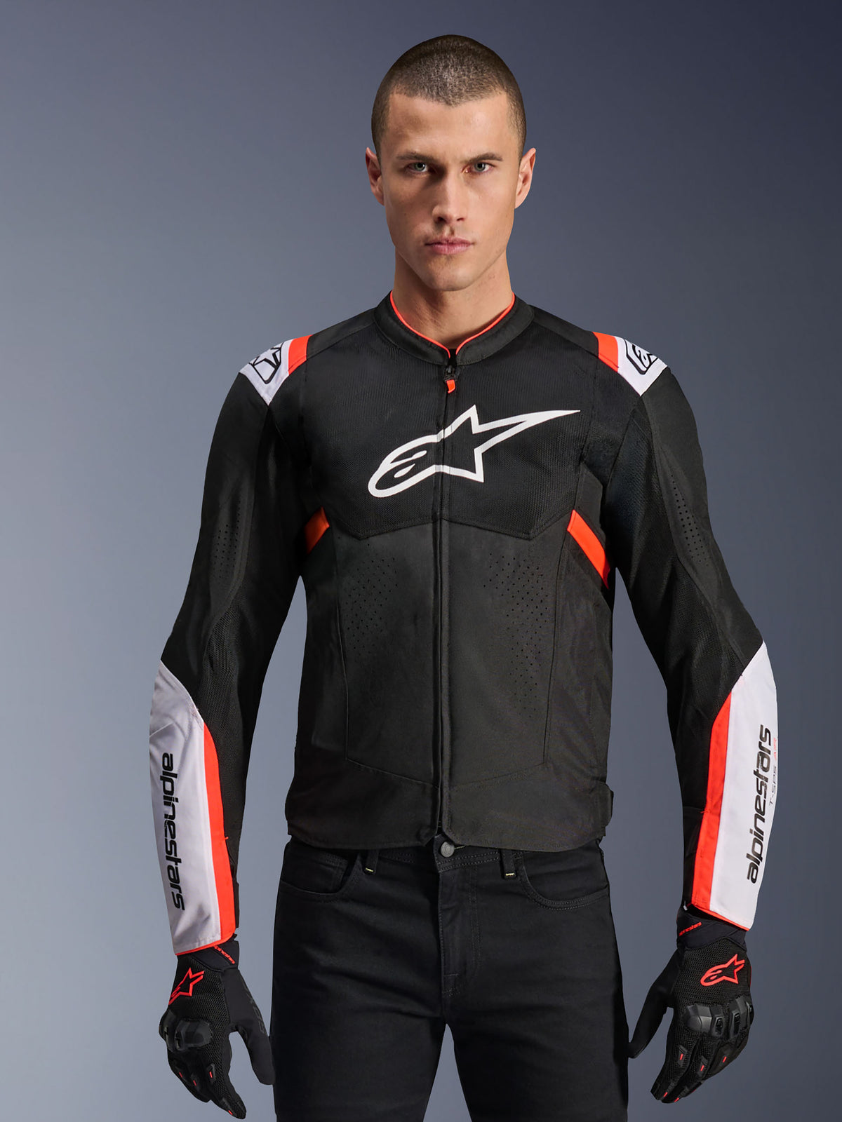 Alpinestars T-Sps Air V2 Jacket, sportieve motorjas, zwart met witte en rood fluo accenten, man draagt een geventileerd mesh-ontwerp met groot logo op de borst en merknaam op de mouwen, gecombineerd met zwarte handschoenen en broek
