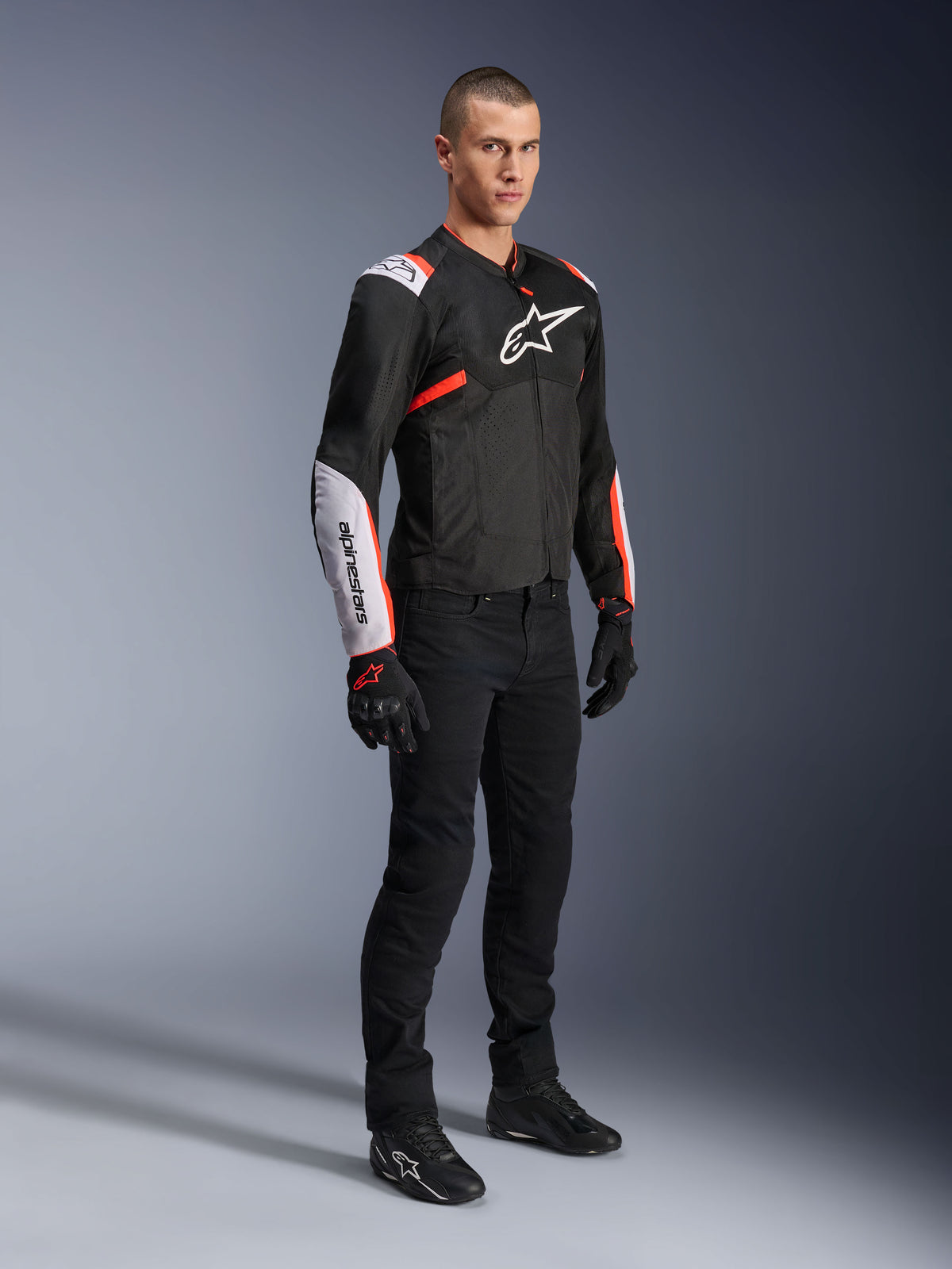 Alpinestars T-Sps Air V2 Jas, Black White Red Fluo, gedragen door een mannelijk model met zwarte motorbroek en handschoenen, textiele motorjas met grote mesh-panelen, witte schouder- en onderarmgedeelten en een opvallend Alpinestars logo op de borst.