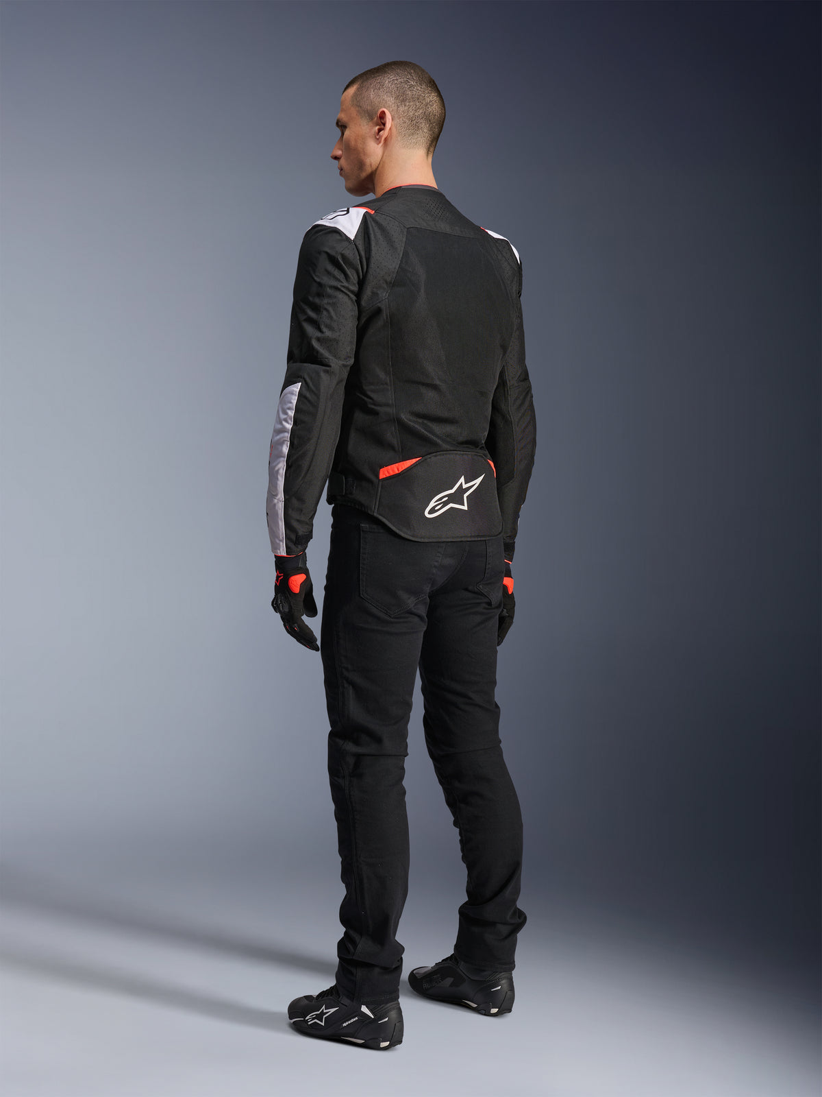 Alpinestars T-Sps Air V2 Jas, textiel motorjas, zwart, wit en fluorood, achteraanzicht op model, mesh-panelen voor ventilatie, wit Astars-logo op de onderrug, gecombineerd met zwarte motorbroek en handschoenen