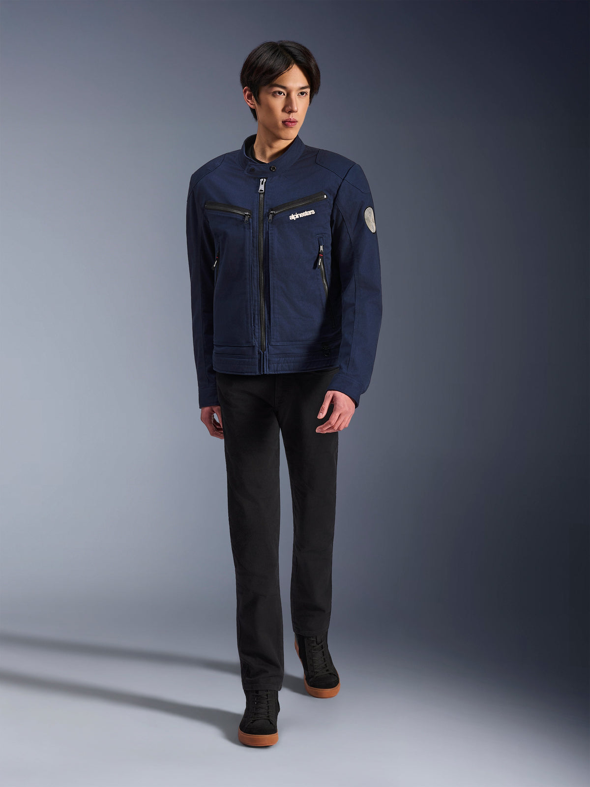 Mannelijk model draagt de Alpinestars Bruiser Jacket in Dark Blue, urban textiele motorjas, gecombineerd met een zwarte broek en zwarte sneakers, werkkleding-geïnspireerde esthetiek met ritszakken en een ronde mouwpatch, eigentijdse streetwear voor motorrijden.