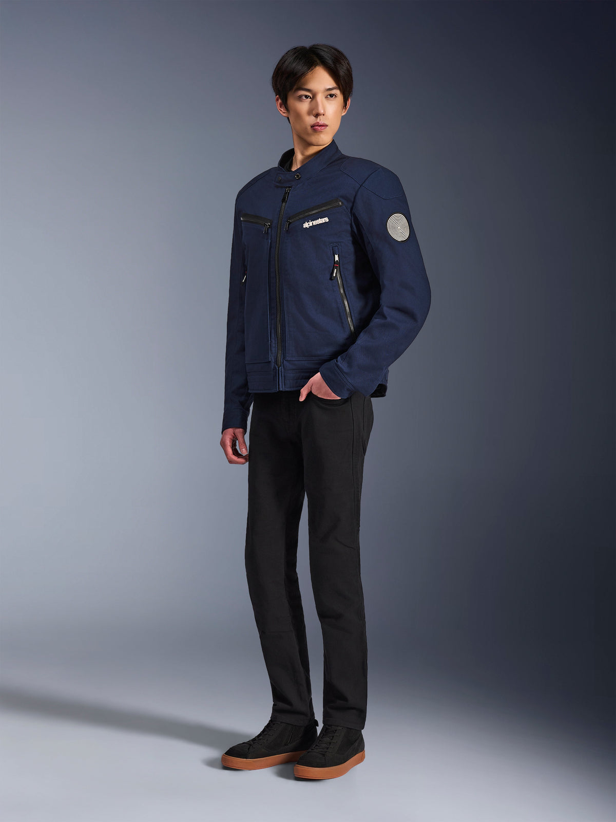 Alpinestars Bruiser Jacket in Dark Blue, urban textiele motorjas met een op werkkleding geïnspireerd ontwerp, voorzien van borstzakken met rits en een Oscar spiraalvormige patch op de mouw, gedragen met een zwarte broek en casual sneakers.