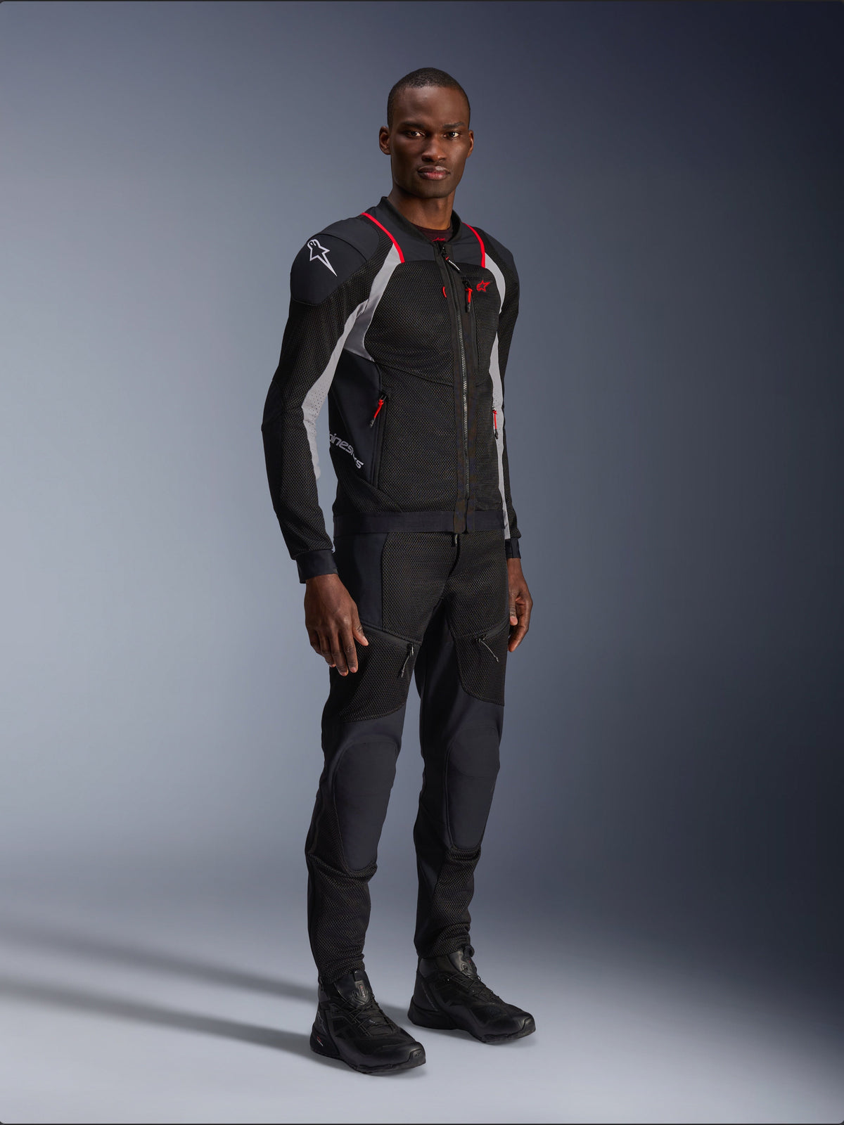 Alpinestars Troop-Air Pro Jacket, Black Ash Gray en Bright Red, technisch gepantserde mesh motorjas, voorzien van rode accenten en een wit logo op de schouder, gedragen met bijpassende zwarte broek en laarzen, moderne urban motorkleding