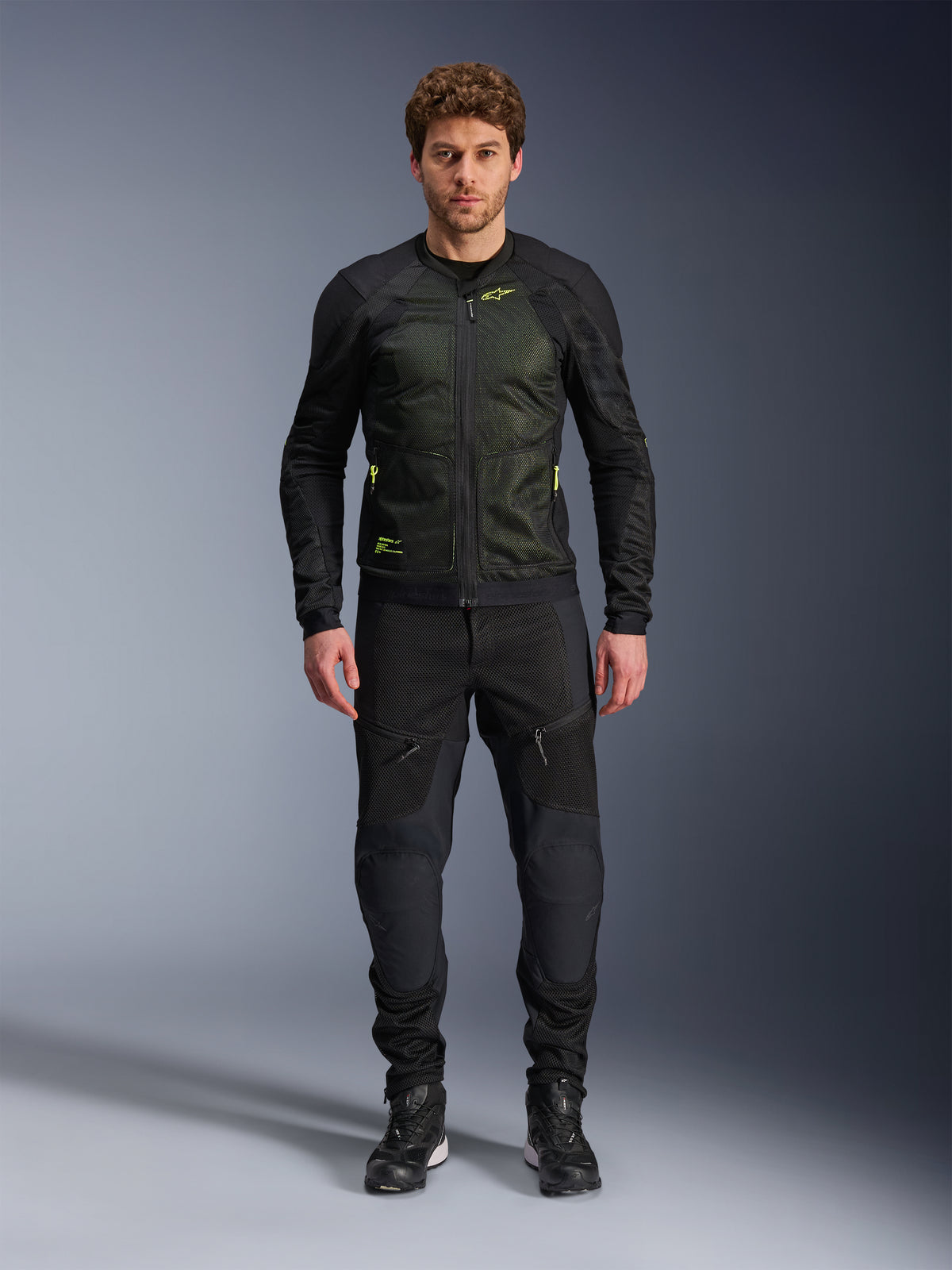Mannelijk model draagt Alpinestars Troop-Air Jacket in Black Yellow Fluo, voorzien van technische mesh-panelen en felgele accenten, gecombineerd met een zwarte motorbroek en schoenen, eigentijdse motorkleding voor warm weer