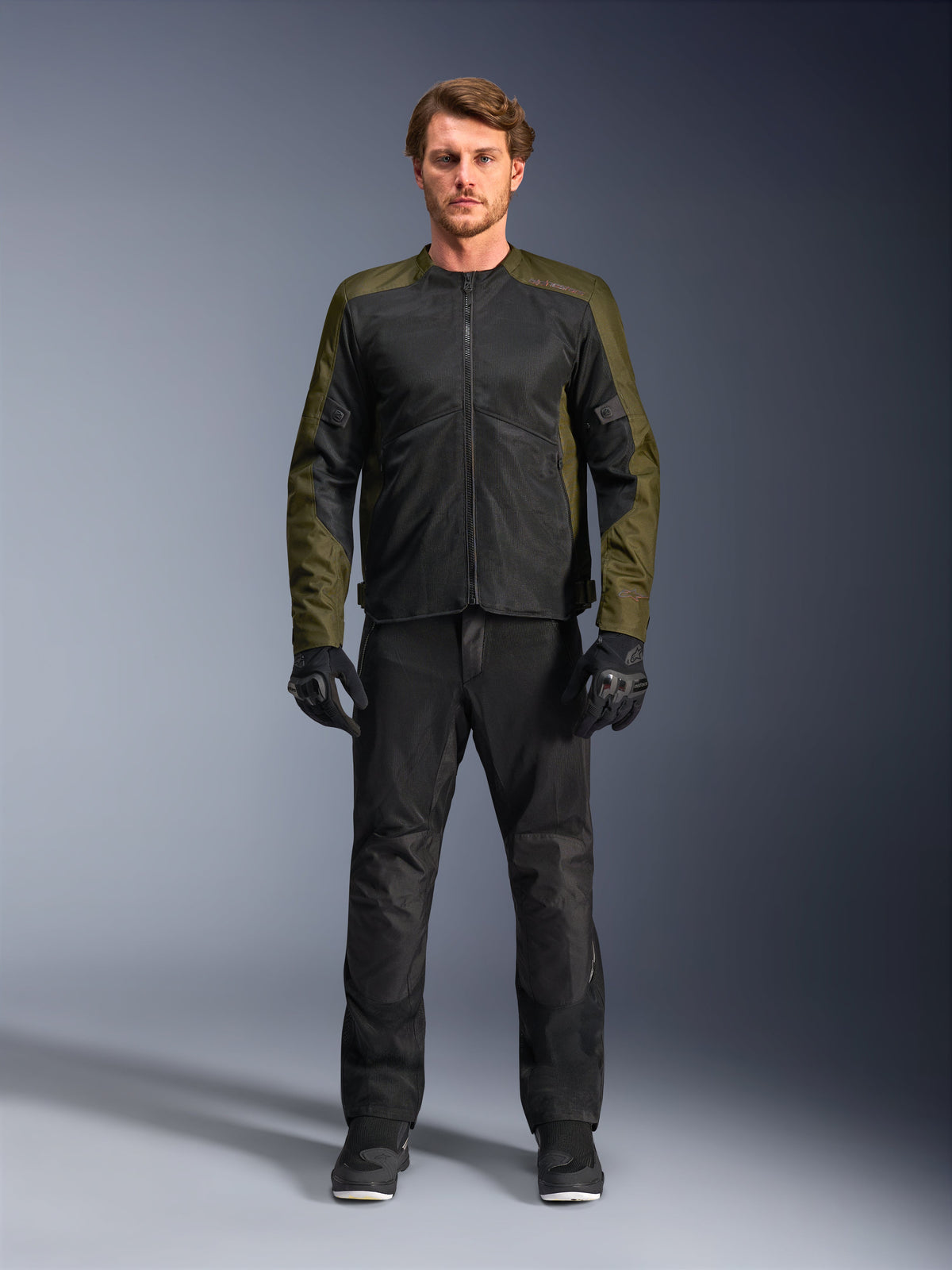 Alpinestars C-1 Air Jas, khaki en zwart, gedragen door een mannelijk model met een zwarte motorbroek en handschoenen, lichtgewicht textielontwerp met tech mesh-panelen, moderne stedelijke motoruitrusting