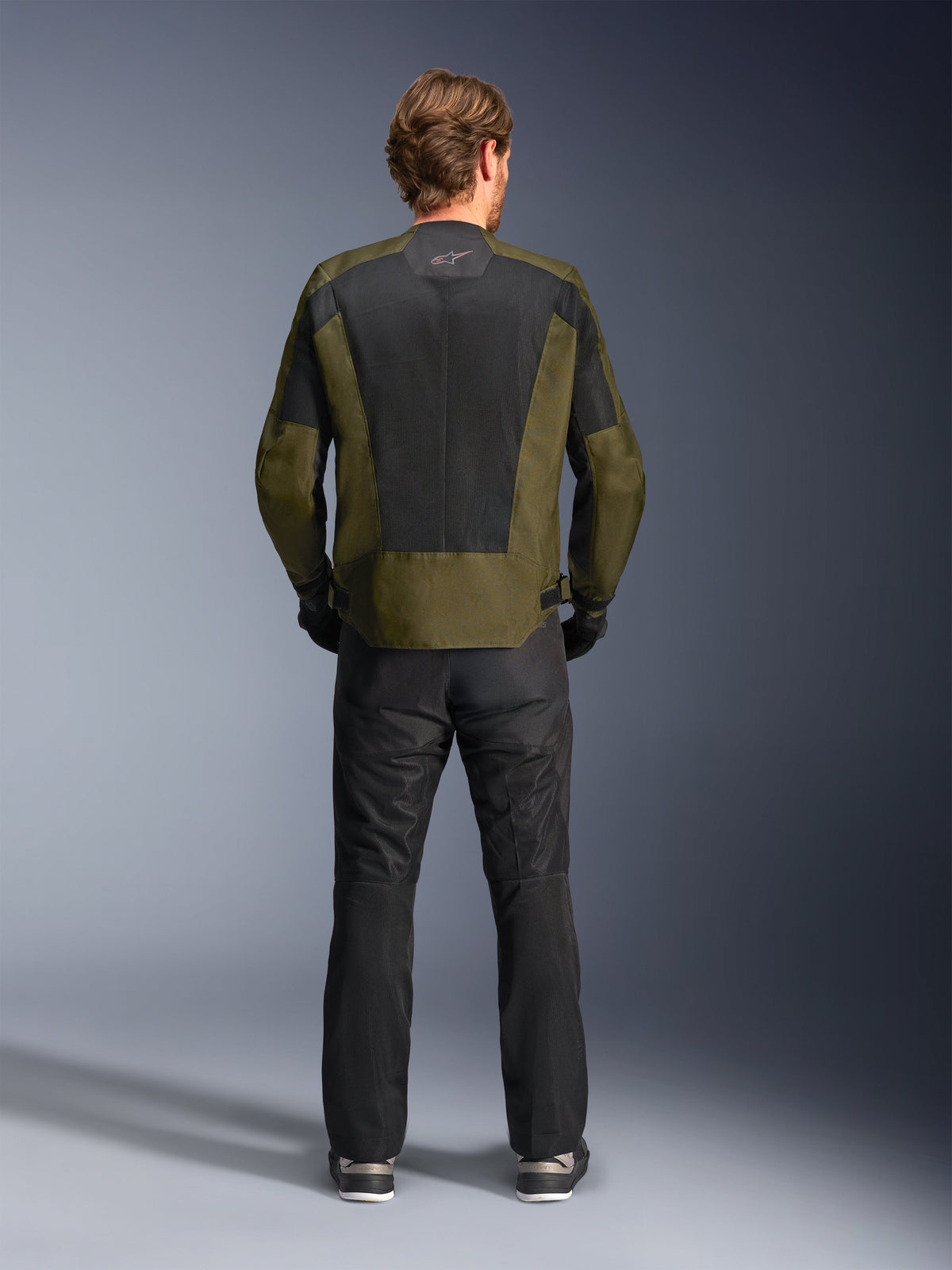 Achteraanzicht van een motorrijder die het Alpinestars C-1 Air Jacket in Khaki en Zwart draagt, voorzien van een groot tech mesh rugpaneel voor ventilatie, gecombineerd met een zwarte motorbroek, urban eigentijdse motorkleding.