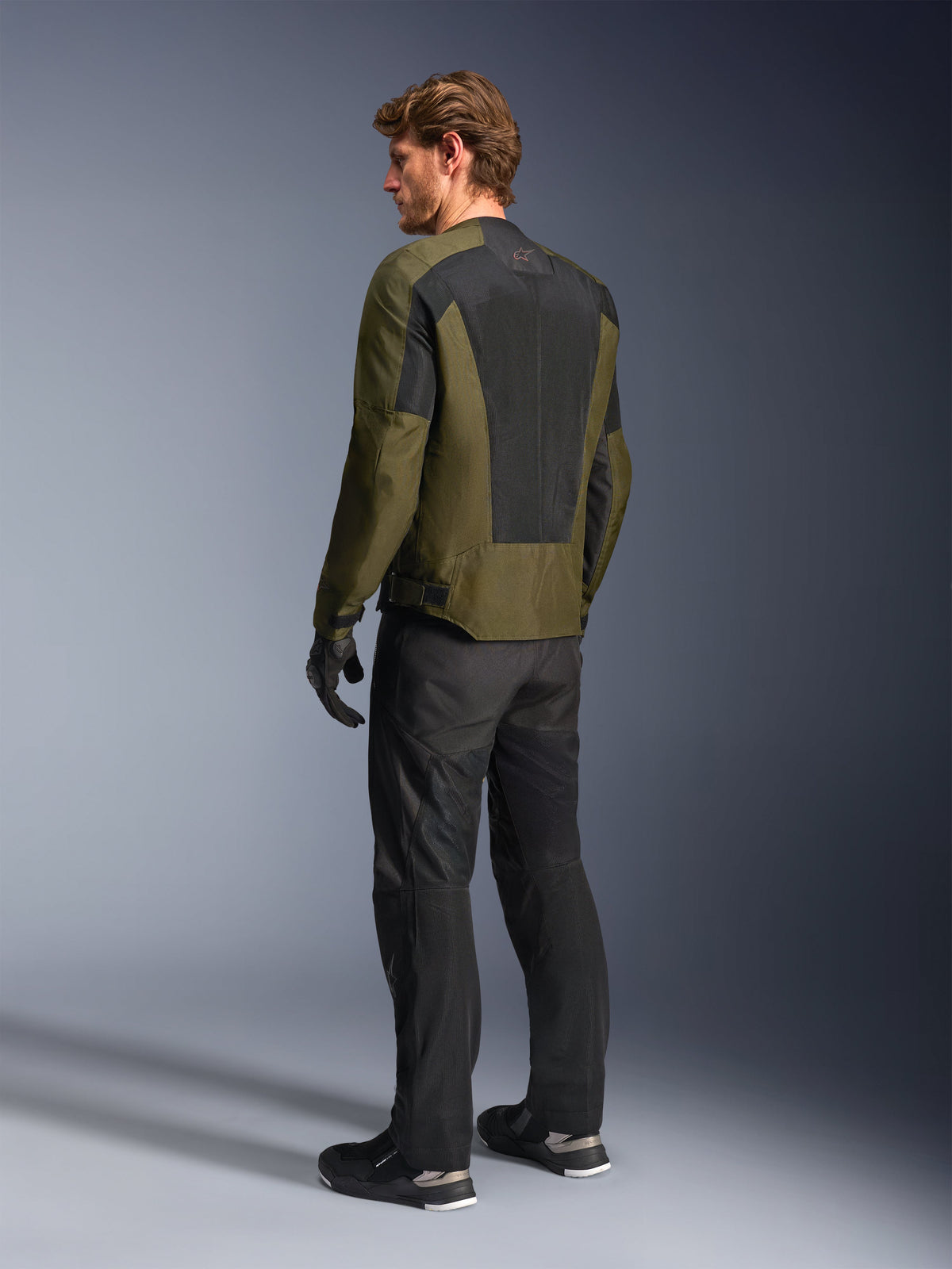 Mannelijk model draagt Alpinestars C-1 Air Jacket in Khaki, urban motorjas, achteraanzicht met nadruk op de grote zwarte mesh-panelen op de rug, gecombineerd met een zwarte technische motorbroek en handschoenen, eigentijdse motoruitrusting voor warm weer