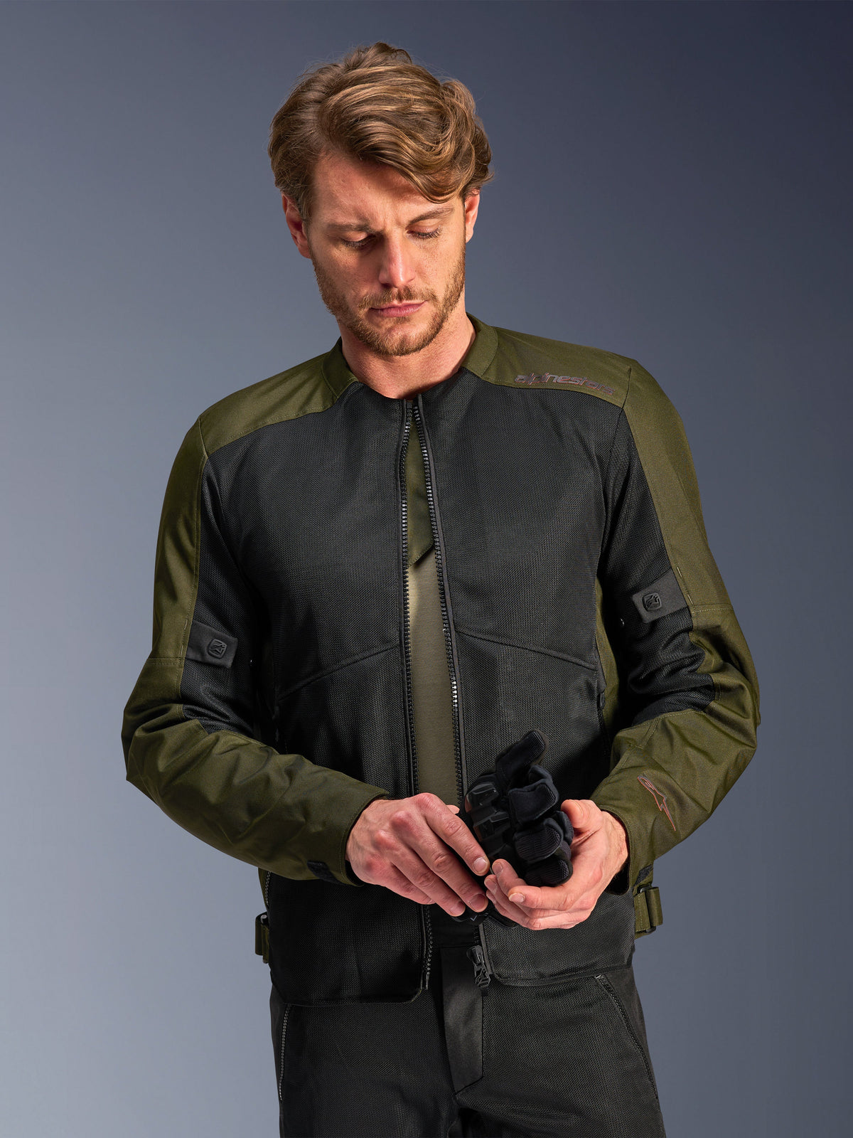 Man die de Alpinestars C-1 Air Jacket in Khaki draagt, urbane motorjas met olijfgroen textiel en zwarte mesh-panelen, kijkt naar beneden terwijl hij zwarte motorhandschoenen vasthoudt, strakke en eigentijdse stijl voor rijden op de weg.