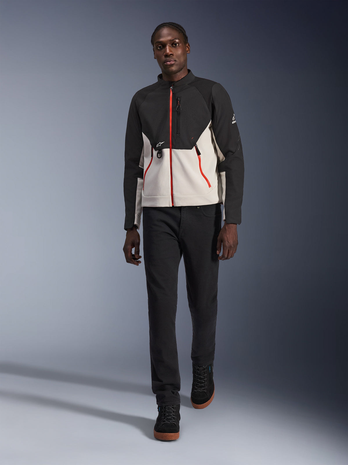 Mannelijk model draagt het Alpinestars Provoke Jacket, een stedelijk motorjack in zwart en metaalgrijs met rode ritsaccenten, gecombineerd met een zwarte broek en zwarte hoge schoenen, moderne straatklare beschermende kleding voor stadsgebruik