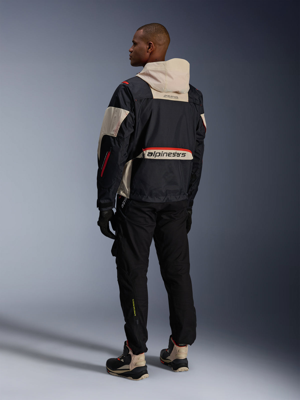 Achteraanzicht van een man die een Alpinestars Acteon broek in het zwart draagt, gecombineerd met een Mogress Air-jas in zwart en crème, zwarte handschoenen en motorlaarzen, eigentijdse urban motoroutfit