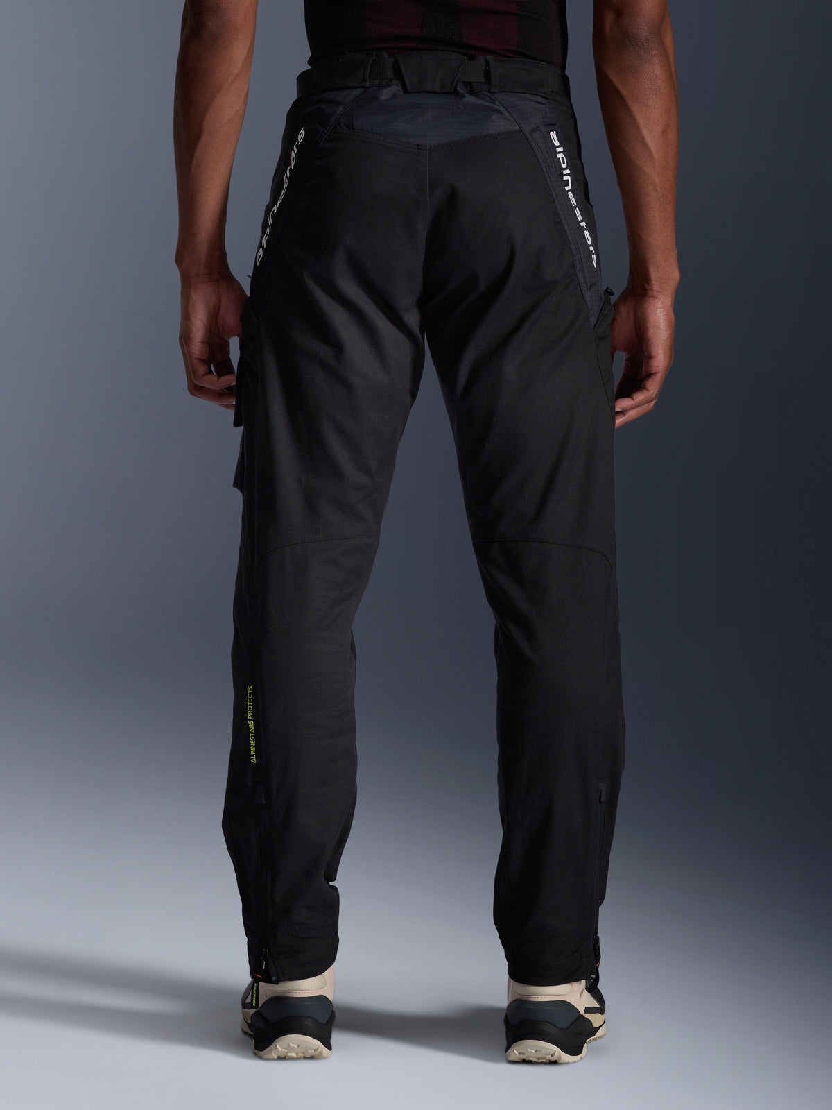 Alpinestars Acteon broek, urban motorbroek, zwart, achteraanzicht met taps toelopende pasvorm, witte merkdetails op de bovenbenen en geel logo op de kuit, verstelbare broekspijpen met ritsen, gedragen met crème sneakers