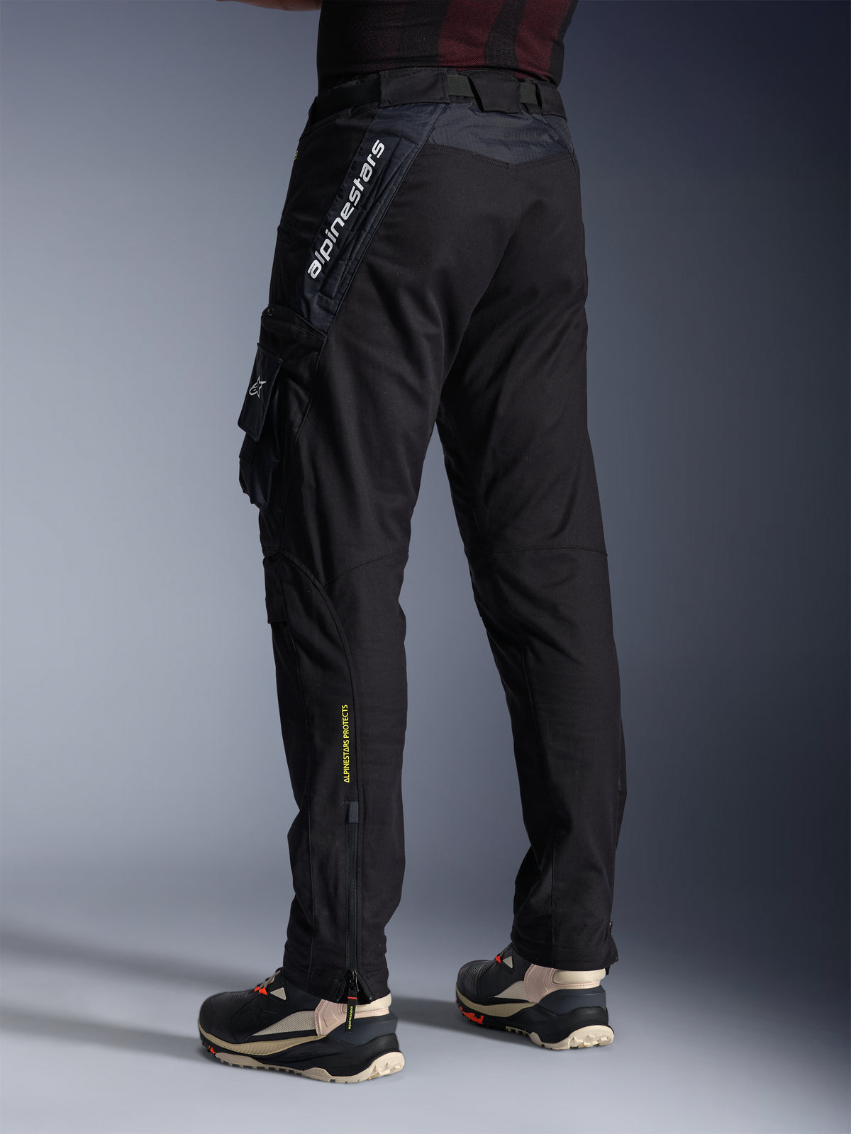 Alpinestars Acteon broek, zwarte motorbroek, achter- en zijaanzicht met een moderne taps toelopende pasvorm, cargozak aan de zijkant met logo, verticale witte Alpinestars-branding op het bovenbeen, gele 'Alpinestars Protects'-tekst op het onderbeen, urban hedendaagse motoruitrusting