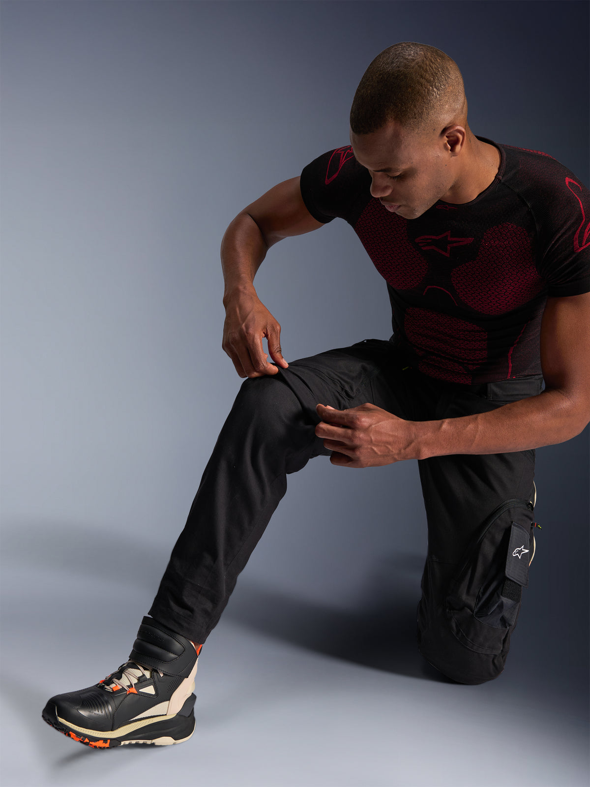 Alpinestars Acteon broek, zwarte urban motorbroek, gedragen door een knielende man die de kniezone aanpast, voorzien van cargozakken en een taps toelopende pasvorm, gecombineerd met een zwart-rode Alpinestars base layer en motorschoenen