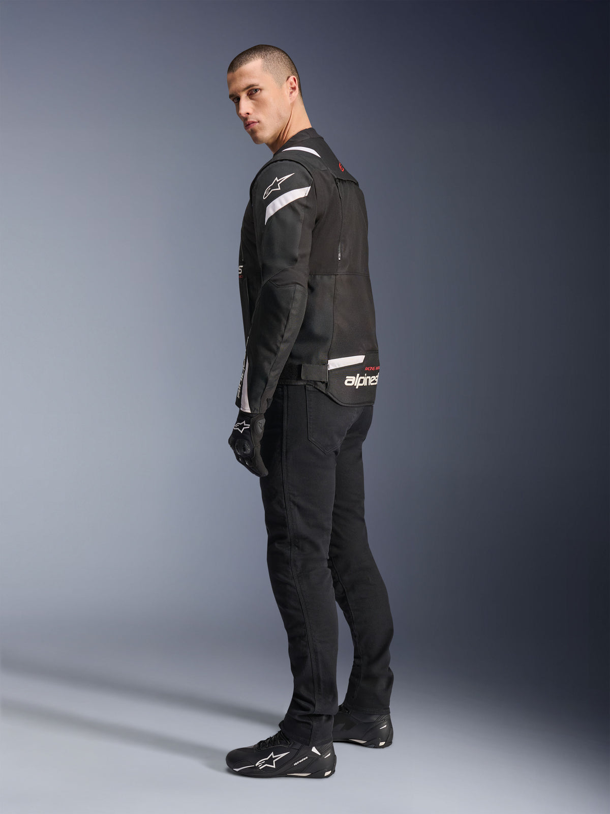 Alpinestars T-Stunt Air Jacket, zwart en wit, modulaire motorjas met tech mesh-panelen, afgebeeld in zij-achteraanzicht op een model, gedragen met zwarte motorbroek en schoenen, witte accenten en branding op de schouders en rug.