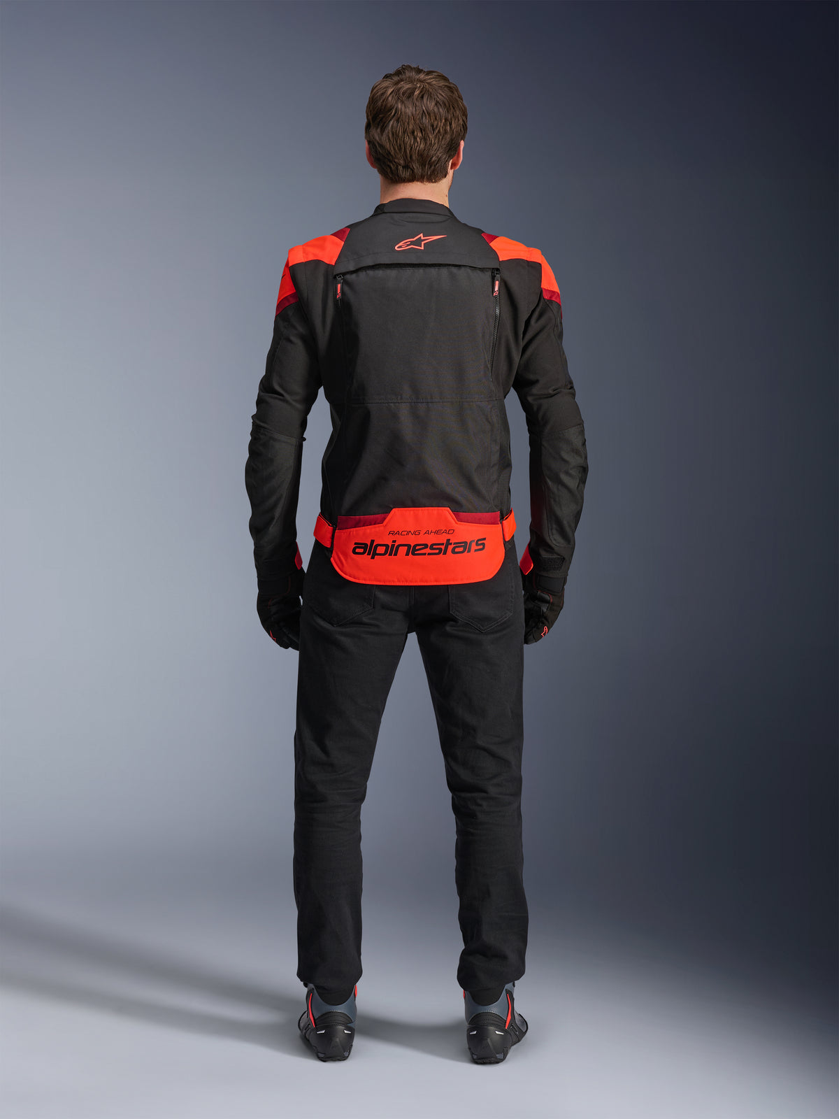 Alpinestars T-Stunt Air Jas, zwart met Red Fluo en Dark Red accenten, achteraanzicht op model, gecombineerd met zwarte motorbroek en laarzen, opvallende Alpinestars-branding op het onderste rugpaneel, modulair ontwerp met mesh-constructie voor ventilatie