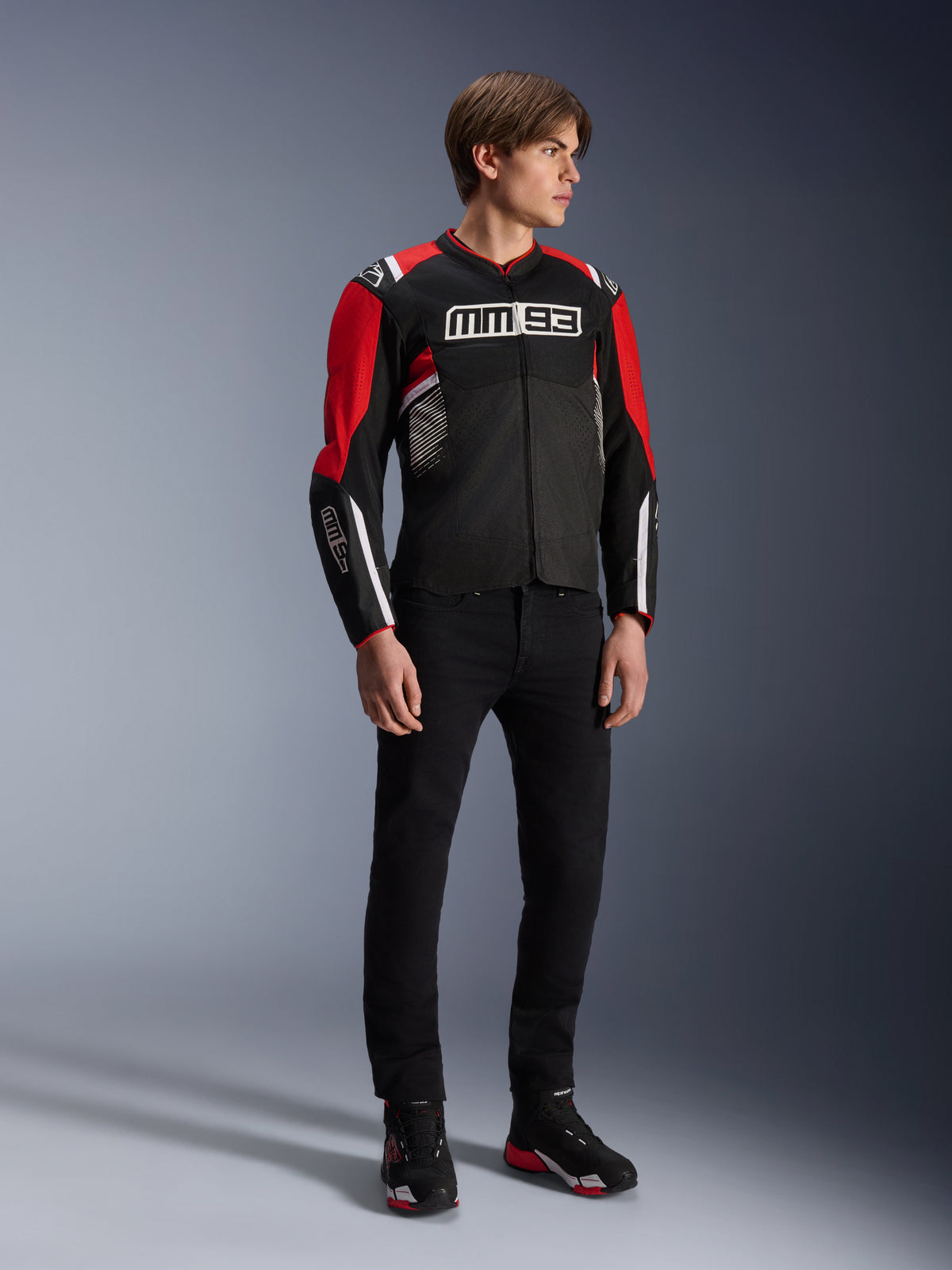 Mannelijk model draagt de Alpinestars MM93 T-SPS Air v2 Jas in Black Bright Red White, met opvallende MM93-branding op de borst en mouwen, gecombineerd met een zwarte broek en motorlaarzen, sportieve urban motorstijl met zichtbare mesh-panelen voor luchtstroom.