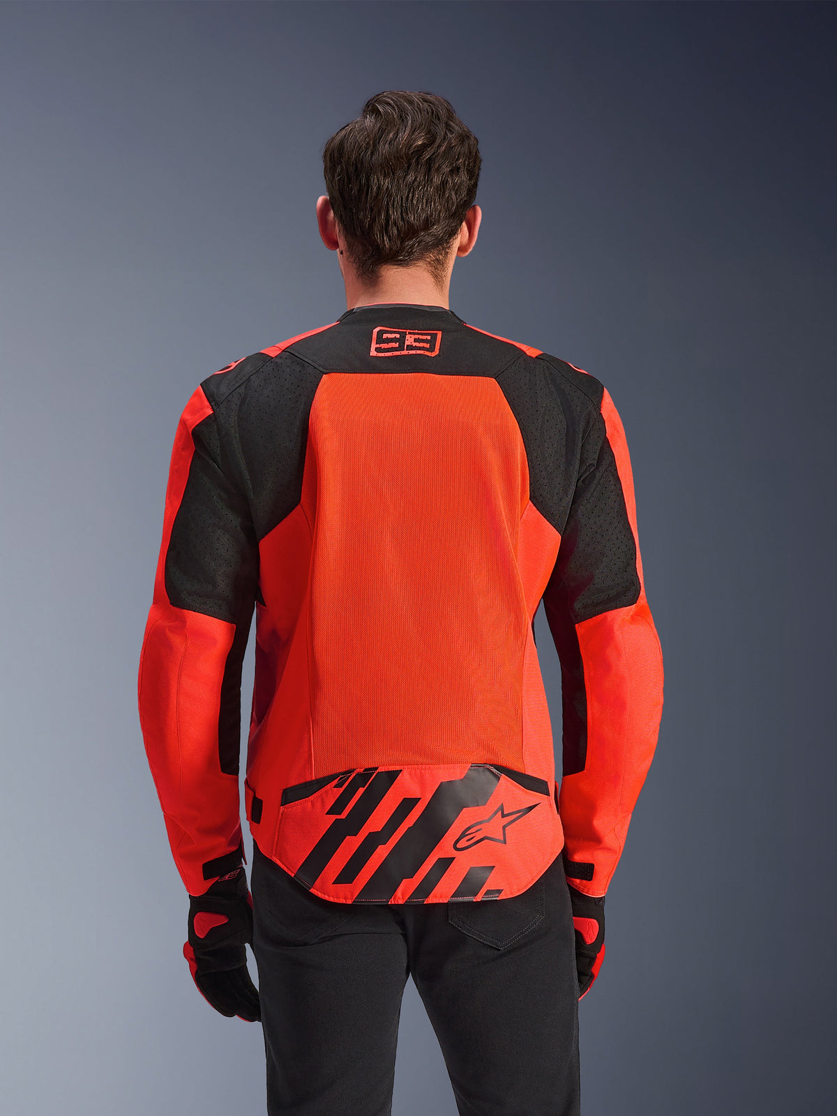 Alpinestars MM93 T-SPS Air V2 Jas, achteraanzicht, fluo rood en zwart, met 93-racelogo op de nek, groot mesh-paneel op de rug voor luchtstroom, achterflap onderaan met Alpinestars-sterlogo en geometrische strepen, gedragen met een zwarte broek en motorhandschoenen
