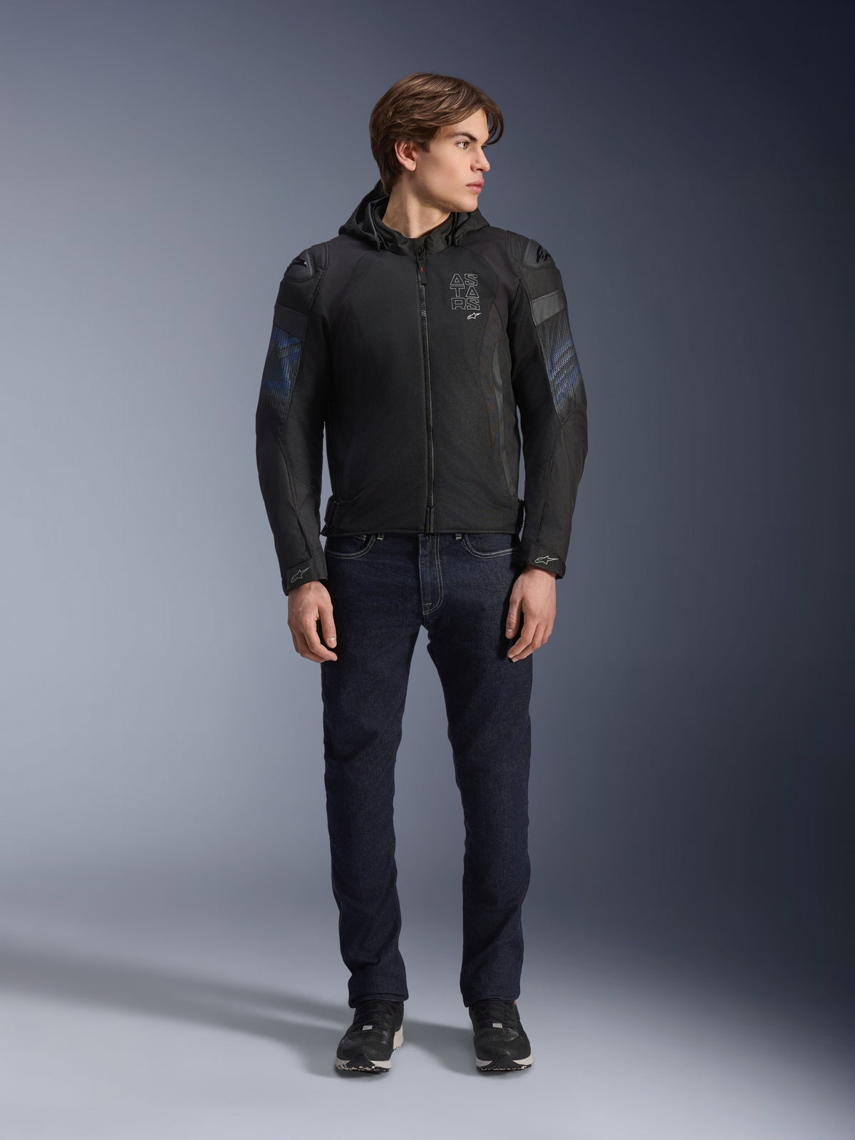 Mannelijk model draagt de Alpinestars Zaca Air Venom WP jas in zwart en elektrisch blauw, textiele motorjas met 3D-mesh panelen en een afneembare capuchon, gecombineerd met een donkere denim jeans, moderne sportgeïnspireerde motoruitrusting