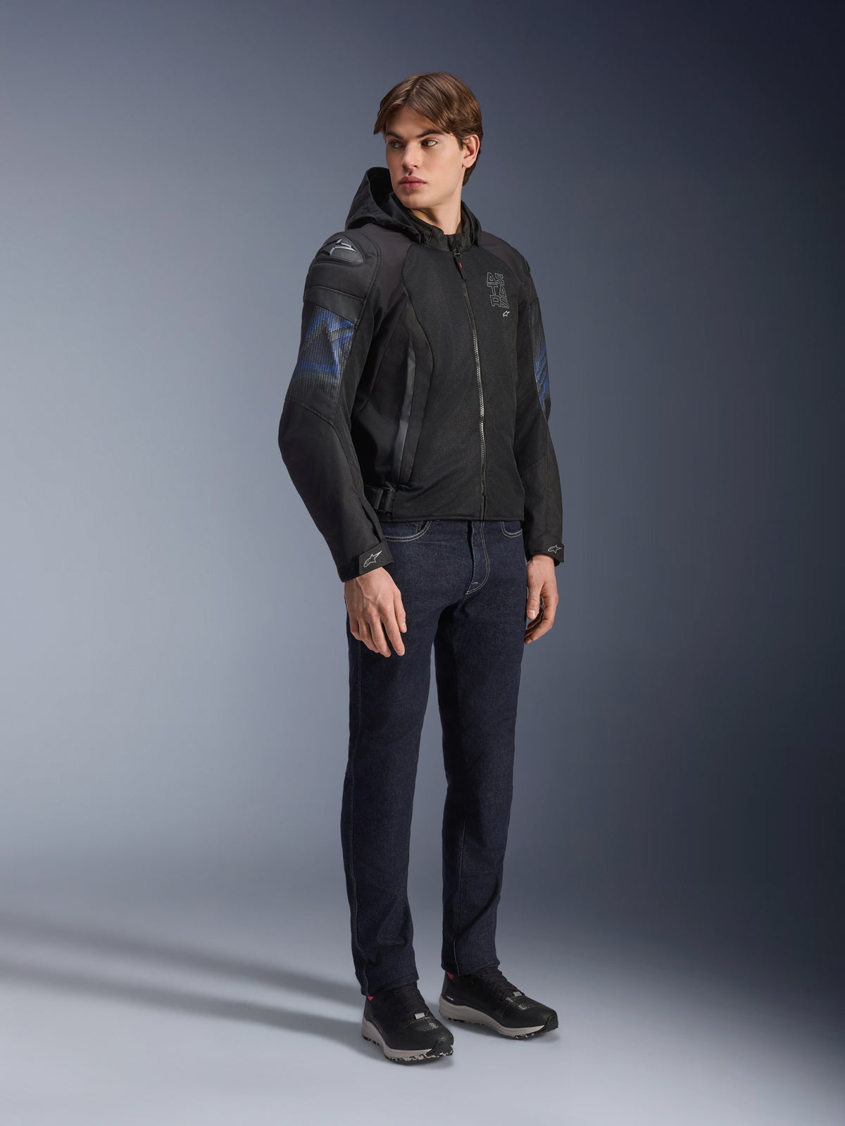 Alpinestars Zaca Air Venom WP Jas, zwarte en elektrisch blauwe textiele motorjas, gedragen door model met donkere jeans en zwarte sneakers, voorzien van een afneembare capuchon, mesh ventilatiepanelen en DFS Lite schouderschuivers, sportieve rijuitrusting voor alle weersomstandigheden.