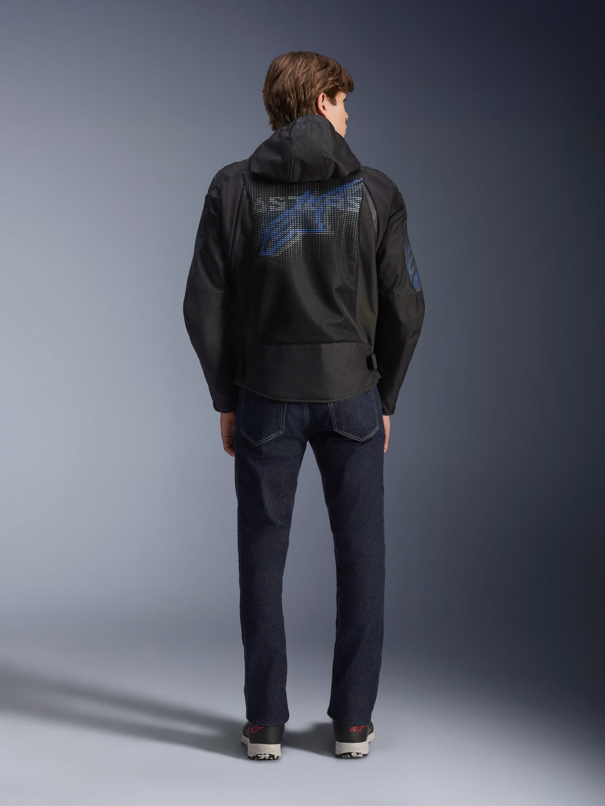 Alpinestars Zaca Air Venom WP Jas, zwart en Electric Blue, achteraanzicht met een groot blauw Alpinestars-logo op het rugpand van mesh, afneembare capuchon, gedragen met donkere denim jeans en sneakers, sportieve all-weather motorkleding.