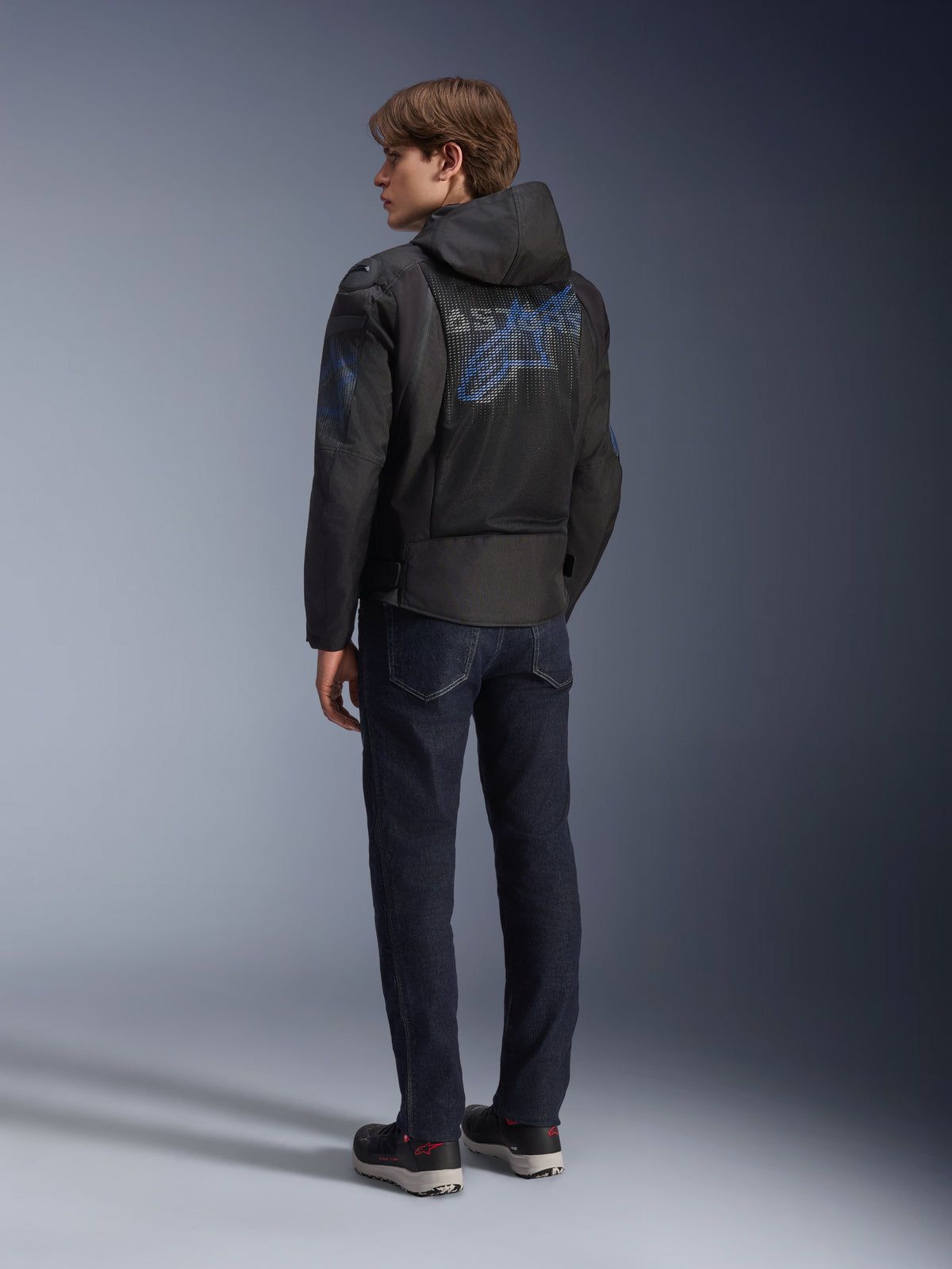 Alpinestars Zaca Air Venom WP jas, zwart en elektrisch blauw, achteraanzicht op een model, mesh textiel motorjas met blauw Astar-logo op de rug, voorzien van een afneembare capuchon, gedragen met donkere jeans en zwarte motorschoenen.