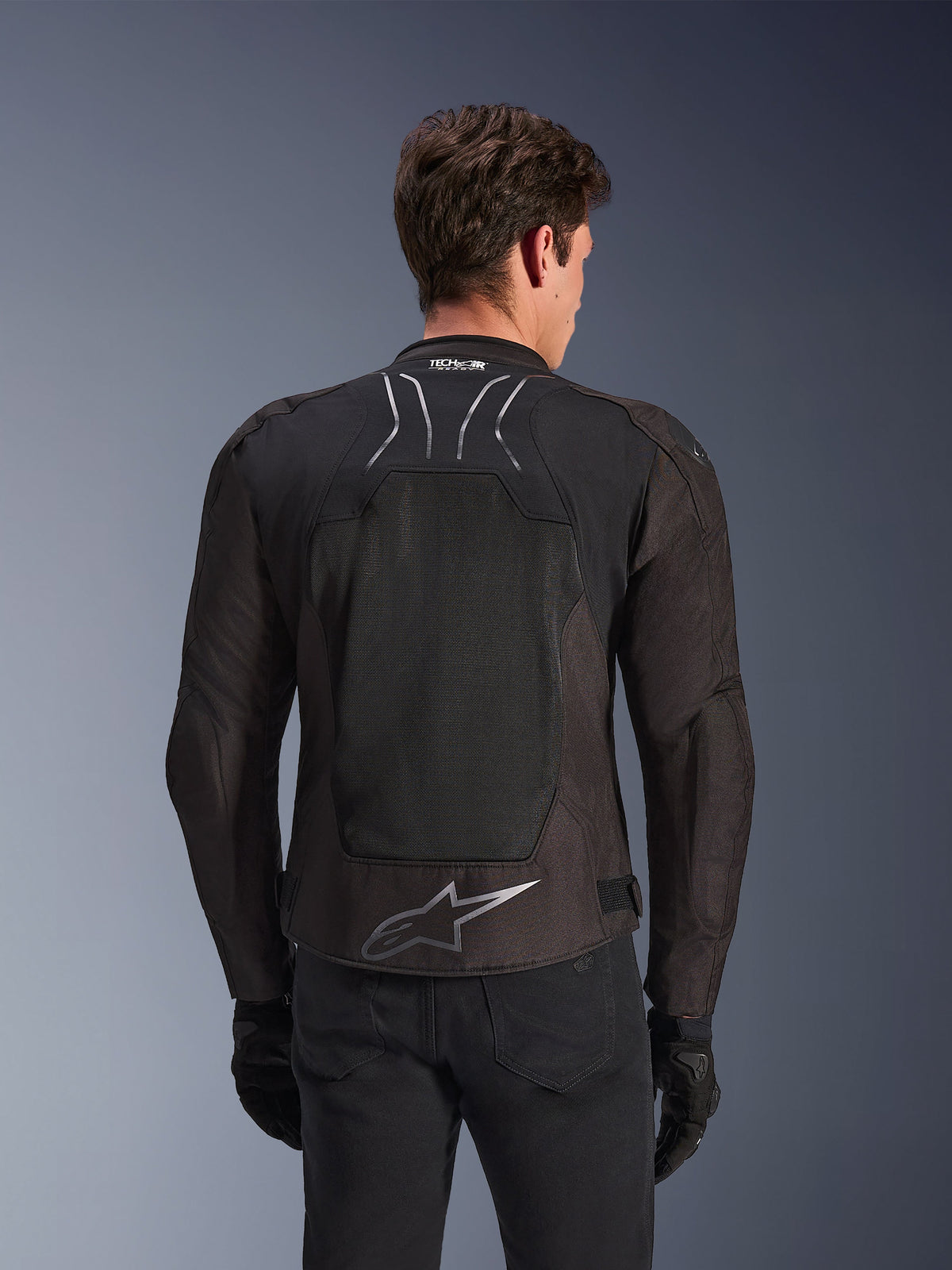 Alpinestars SMX Air Jas in Black Anthracite, achteraanzicht op een model, voorzien van een groot tech mesh rugpaneel voor ventilatie, Tech-Air Ready-branding op de kraag en een zilveren Astar-logo op de onderrug, gecombineerd met zwarte handschoenen en een motorbroek.