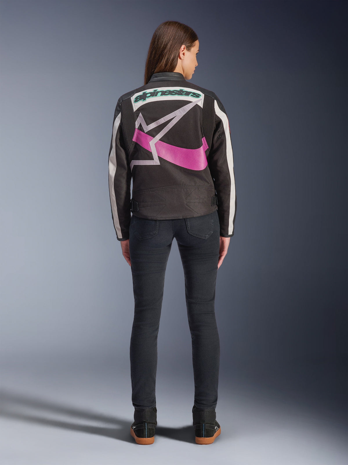 Achteraanzicht van een vrouw met de Alpinestars Idles jas, zwart met gebroken witte strepen, voorzien van een groot paars en wit Alpinestars ster-logo en groen woordmerk op de rug, gecombineerd met donkere slim-fit jeans, vintage-geïnspireerde stedelijke motorkleding