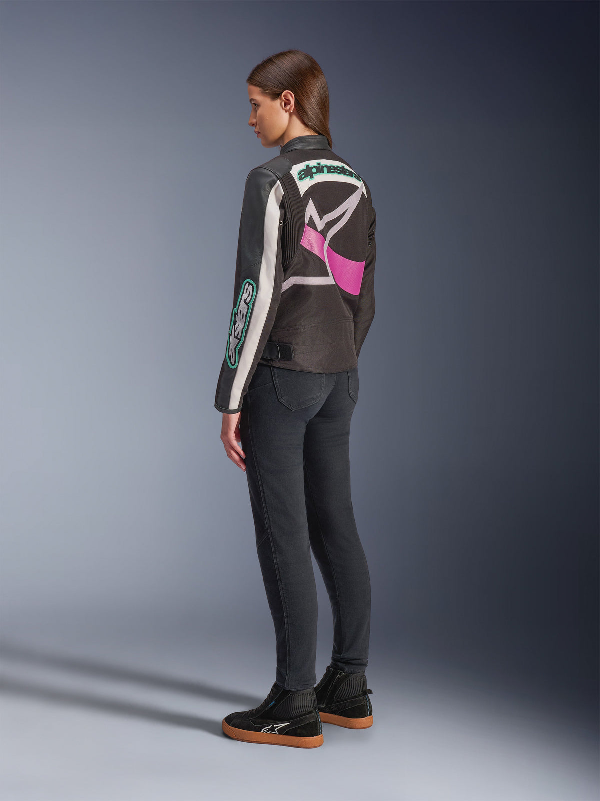 Alpinestars Idles damesjas, motorjas, zwart, off-white en paars, achteraanzicht op model, met een groot rondom lopend Alpinestars-logo en vintage racing-geïnspireerd ontwerp, gecombineerd met een donkere jeans en zwarte schoenen.