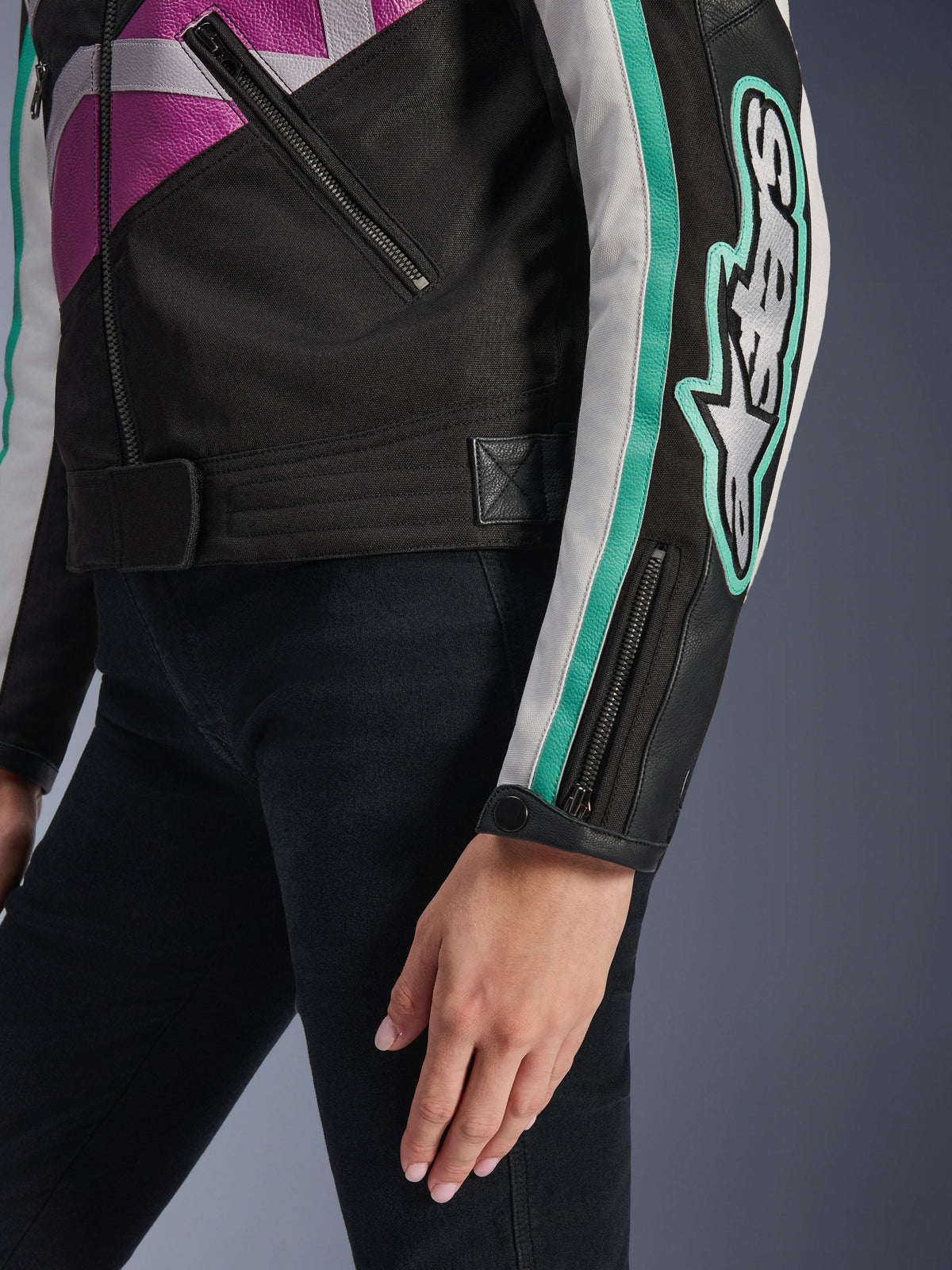 Alpinestars Idles damesjas, close-up van de mouw en zijkant van de romp, zwart met paarse, witte en teal accenten, voorzien van een opvallend Alpinestars-logo op de arm, manchetten met rits en drukknoop en een verstelband in de taille, vintage-geïnspireerde motorkleding.