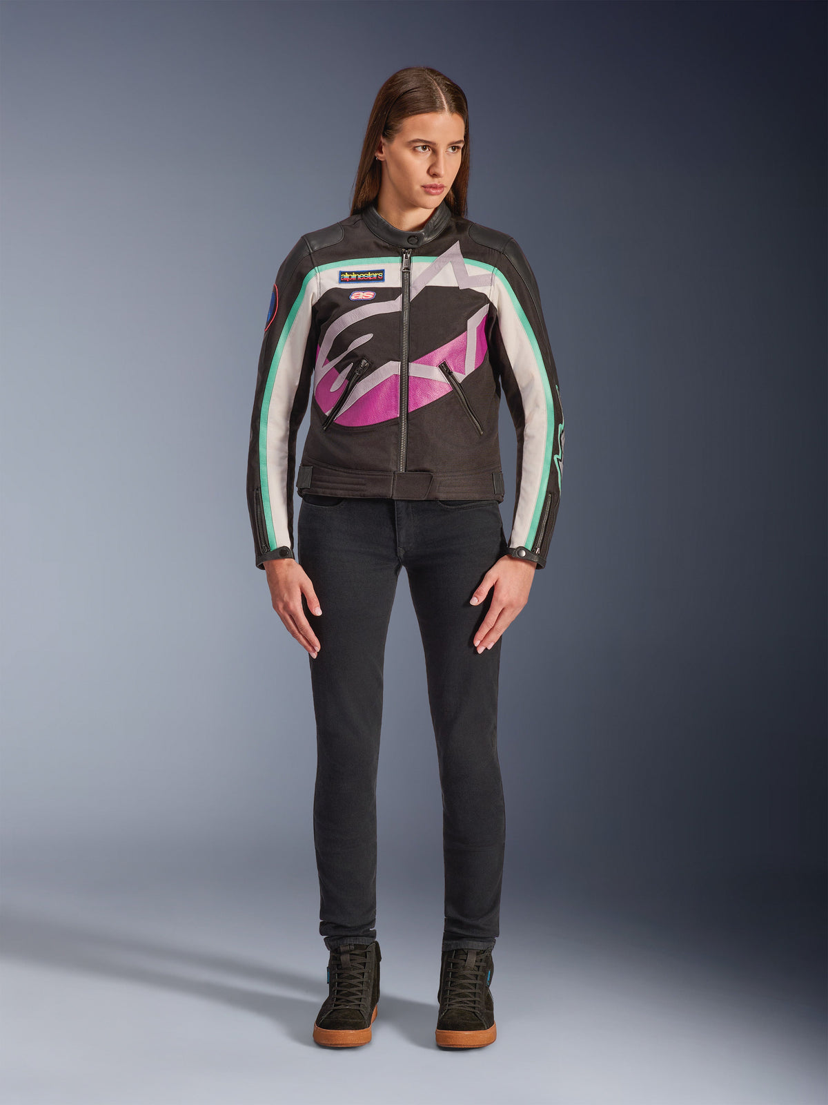 Vrouw draagt Alpinestars Idles damesjas, zwart, off-white en paars, vintage racing-geïnspireerd ontwerp met groot rondom lopend logo en teal accenten, gestyled met een zwarte jeans en motorsneakers, urban motorkleding