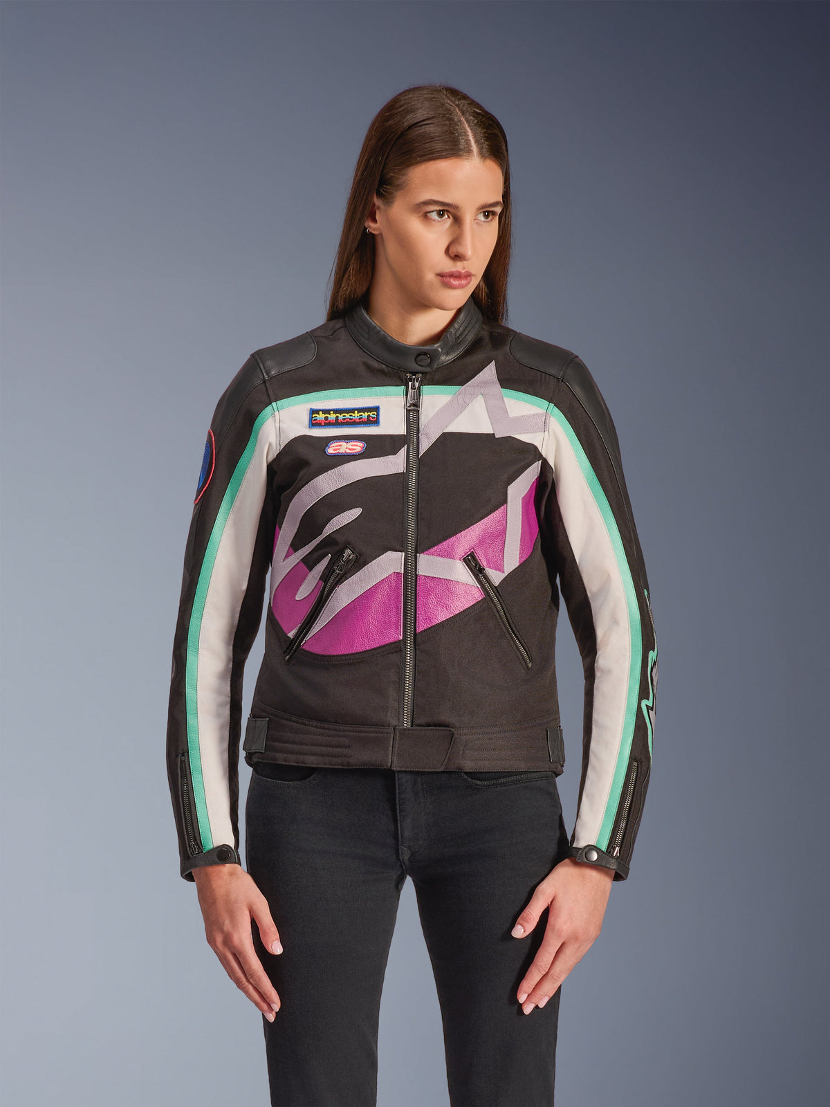 Alpinestars Idles damesjas, motorjas, zwart met gebroken wit, paars en turquoise kleurvlakken, vintage op racing geïnspireerde stijl met groot abstract logo op de borst, merkpatches en ritssluiting aan de voorzijde, gedragen door model met zwarte jeans