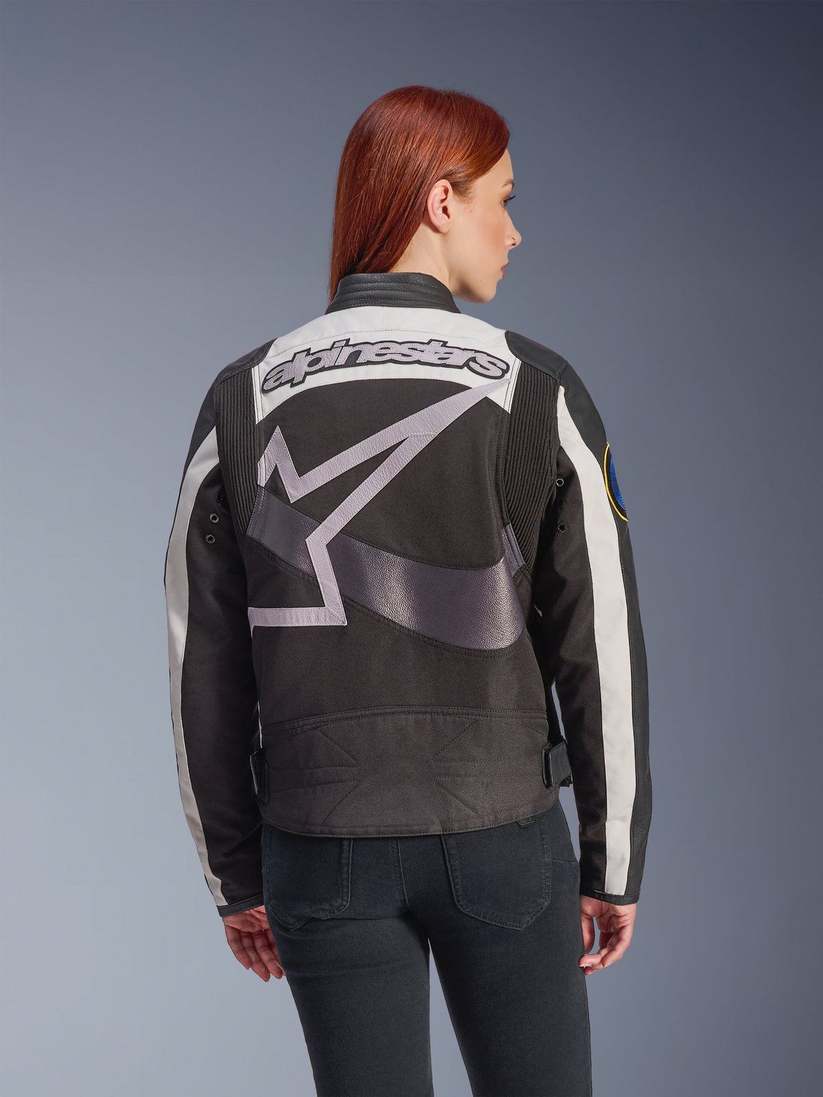 Alpinestars Idles damesjas, zwart en gebroken wit, achteraanzicht met een groot grijs sterlogo en Alpinestars-woordmerk, gedragen door een roodharig model, vintage-geïnspireerde urban motorjas met accordeonpanelen.