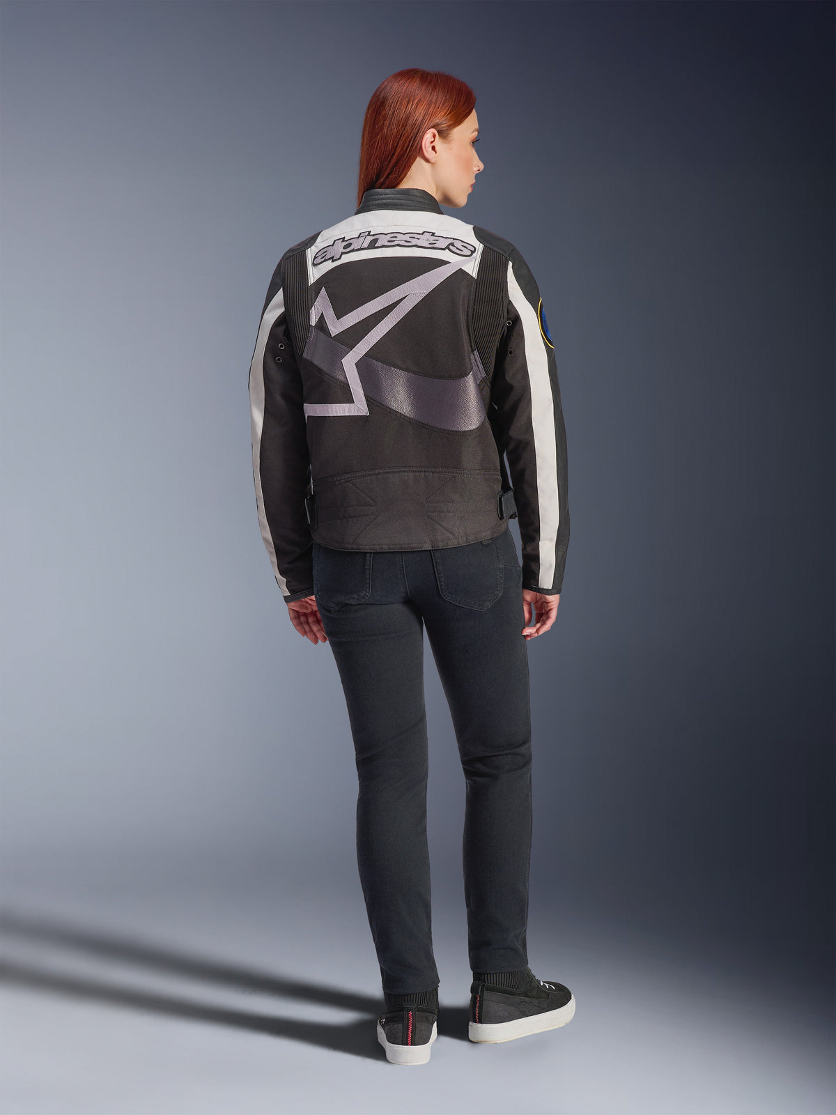 Alpinestars Idles damesjas, urban motorjas, zwart en gebroken wit, achteraanzicht met een groot grijs Alpinestars-logo en woordmerk, gedragen met een zwarte slim-fit jeans en sneakers, vintage-geïnspireerd ontwerp voor stedelijke ritten