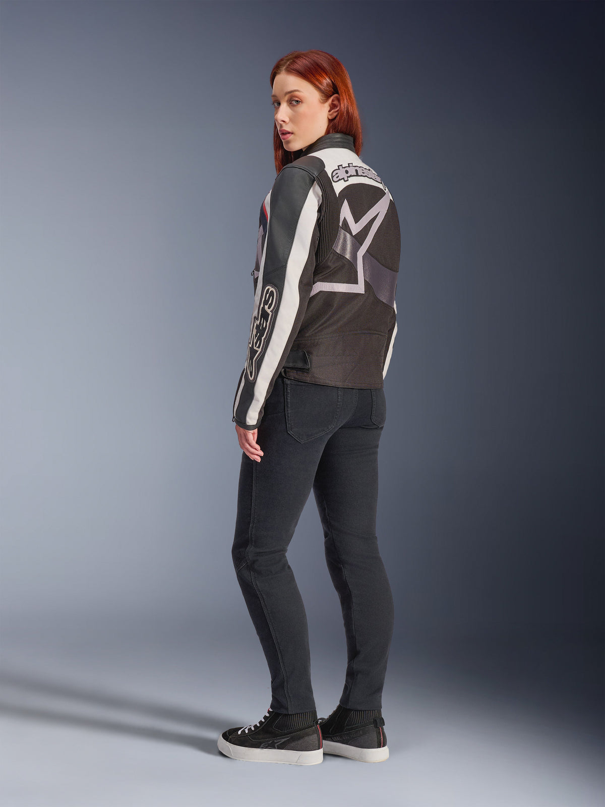 Vrouw in Alpinestars Idles Jacket, achteraanzicht, Off White Flame Red en Black, met een groot sterlogo op de rug en Alpinestars-woordmerk op de mouw, gecombineerd met een zwarte slim-fit broek en sneakers, urban motorkleding voor dames