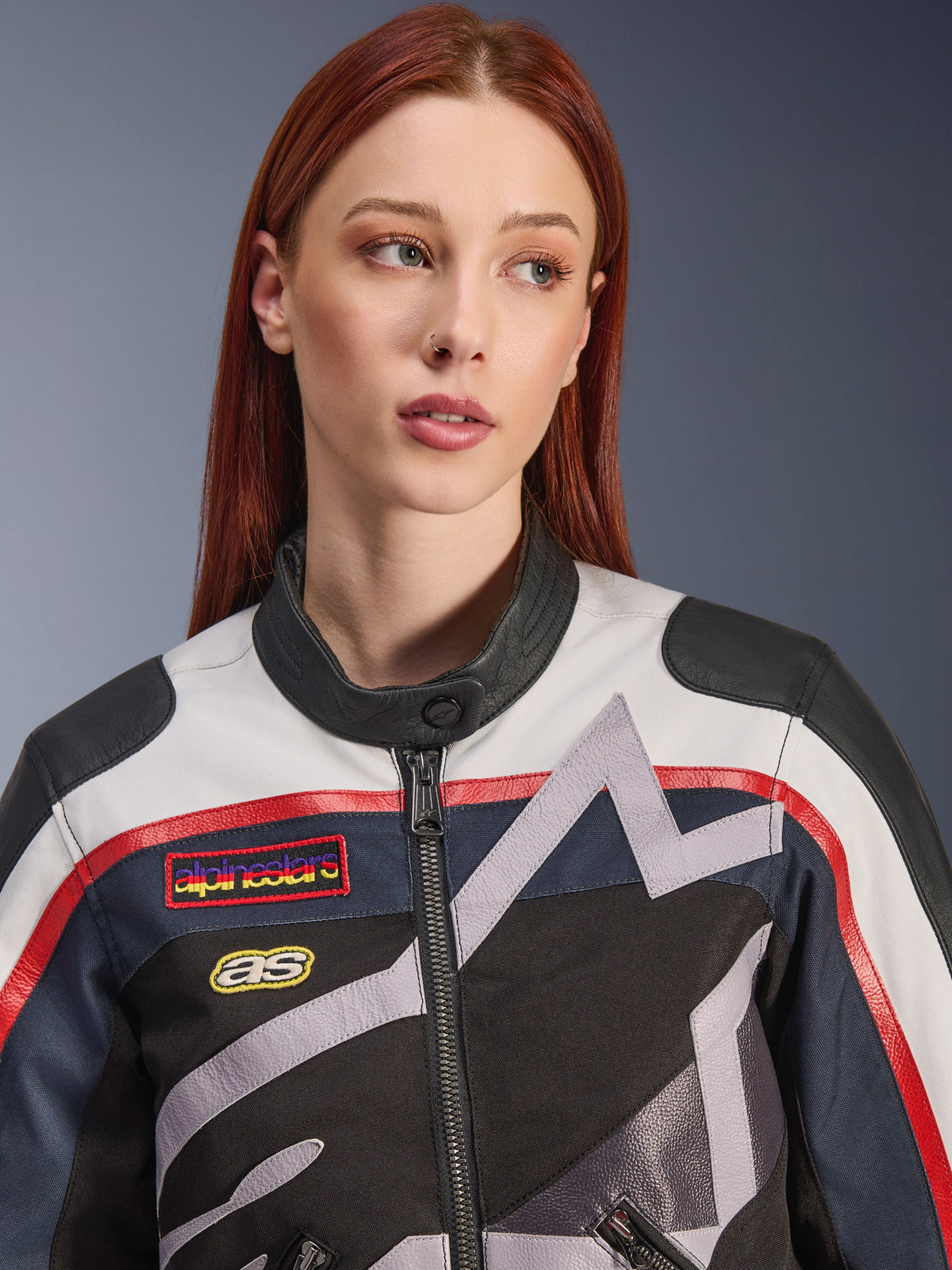 Alpinestars Idles damesjas, close-up van een vrouwelijk model met rood haar, Off White Flame Red en Black kleurstelling, vintage-geïnspireerd racing-ontwerp met logopatches en grijze geometrische ster-graphics, urban motorrijuitrusting.