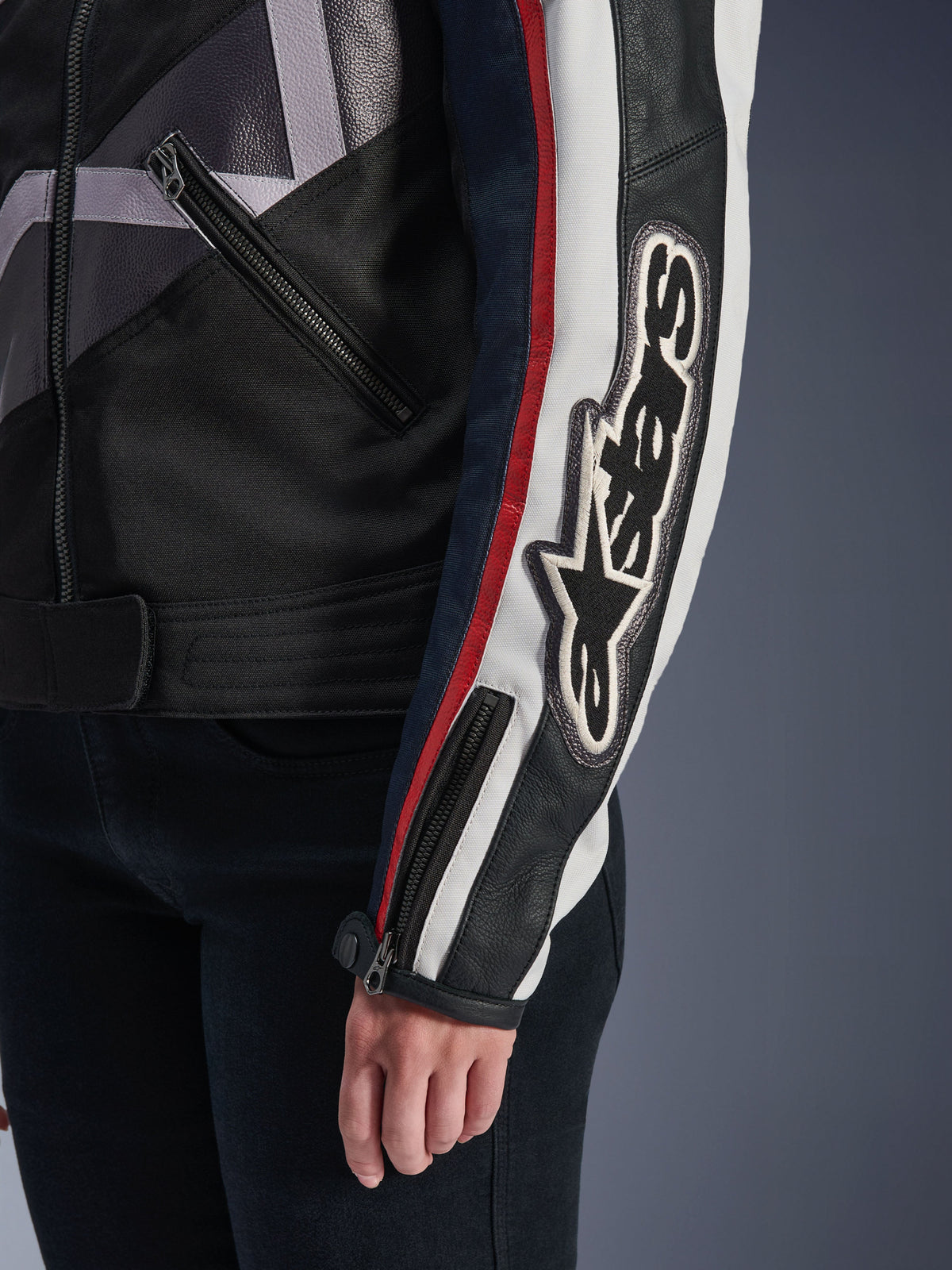 Alpinestars Idles damesjas, urban motorjas, Off White Flame Red Black, close-up van de mouw met een groot geborduurd Alpinestars-logo, rode en witte racestrepen, zwarte textiel- en lederen constructie, en metalen ritsaccenten.