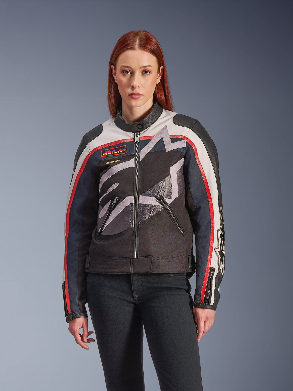 Alpinestars Idles Jacket, damesmotorjas, gebroken wit, vlammend rood en zwart, voorzien van een groot gestileerd sterlogo, vintage-geïnspireerde borstpatches, hoge kraag, zakken met rits en gestreepte mouwen, gedragen door een vrouwelijk model met een donkere jeans